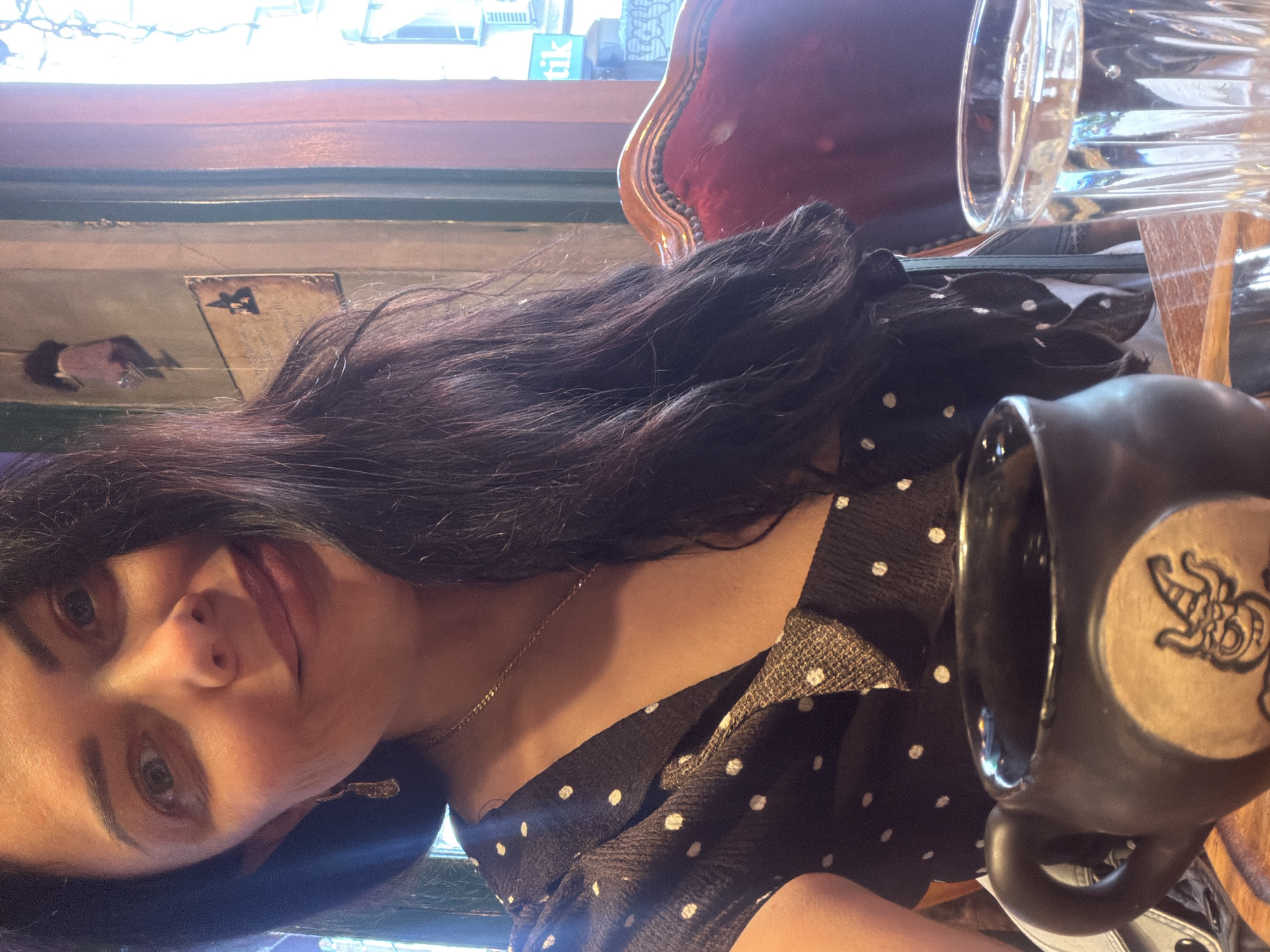 A woman with dark hair and fair skin is seated at a table in a cafe in Kadıköy, Turkey. She is wearing a black and white polka dot dress and a delicate gold chain necklace. She is looking directly at the camera. A black mug with a light-colored emblem on it is prominently displayed in the foreground, positioned near her. A glass of water is visible behind the mug. The cafe has a vintage aesthetic with a dark wood countertop, and a burgundy velvet chair is partially visible in the background. Art or decorative elements adorn the wall in the background. A window with a view of the street outside is visible above.