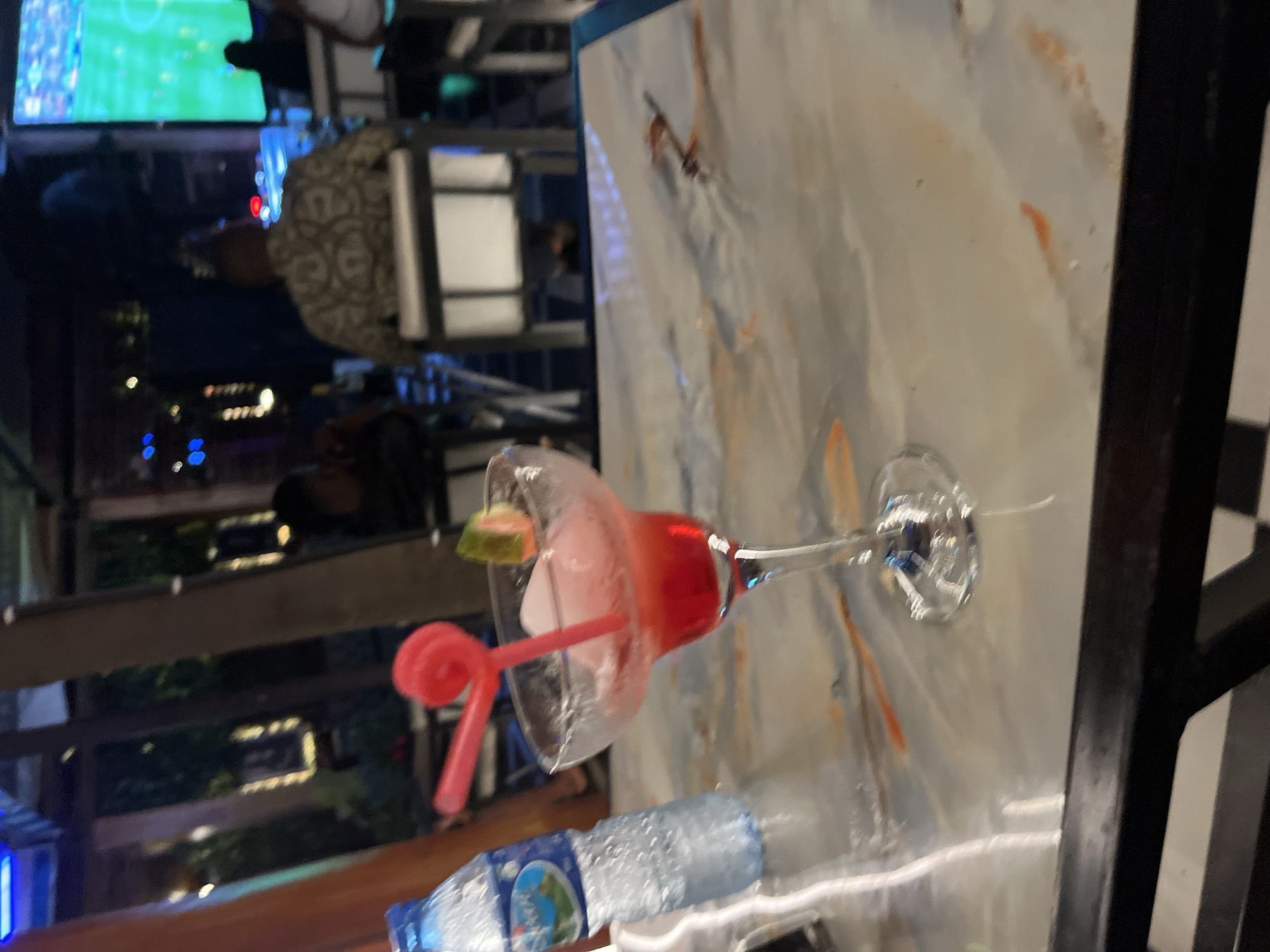 In a dimly lit outdoor seating area, likely at a restaurant or bar in Abuja, Nigeria, a frozen pink beverage sits in a margarita glass on a marble-patterned table. A red swirled straw is inserted into the drink, and a small piece of fruit, possibly watermelon, garnishes the rim. To the left of the drink, a partially visible, condensation-covered bottle of water stands upright. In the background, patrons are seated at tables and a large screen displays a green sports field. The overall ambiance suggests a relaxed evening setting.