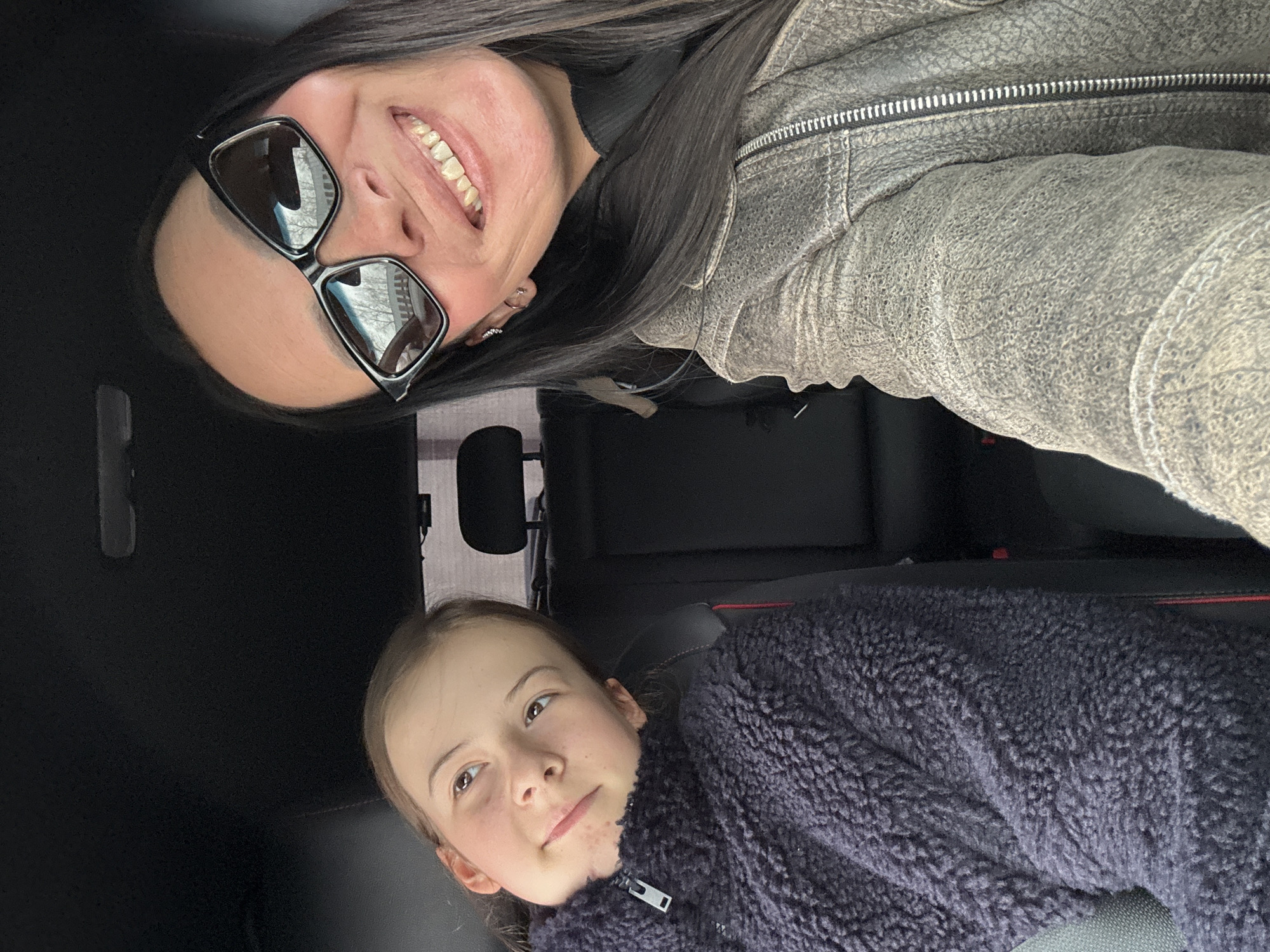 A photo shows an adult woman and a child in what appears to be a car interior. The woman, with dark hair and sunglasses, is wearing a light-colored jacket and smiling. The child is wearing a dark, fuzzy jacket. The background shows black seats and interior car components. The location in the city of Chaikovsky, Russia, is not evident from the image.