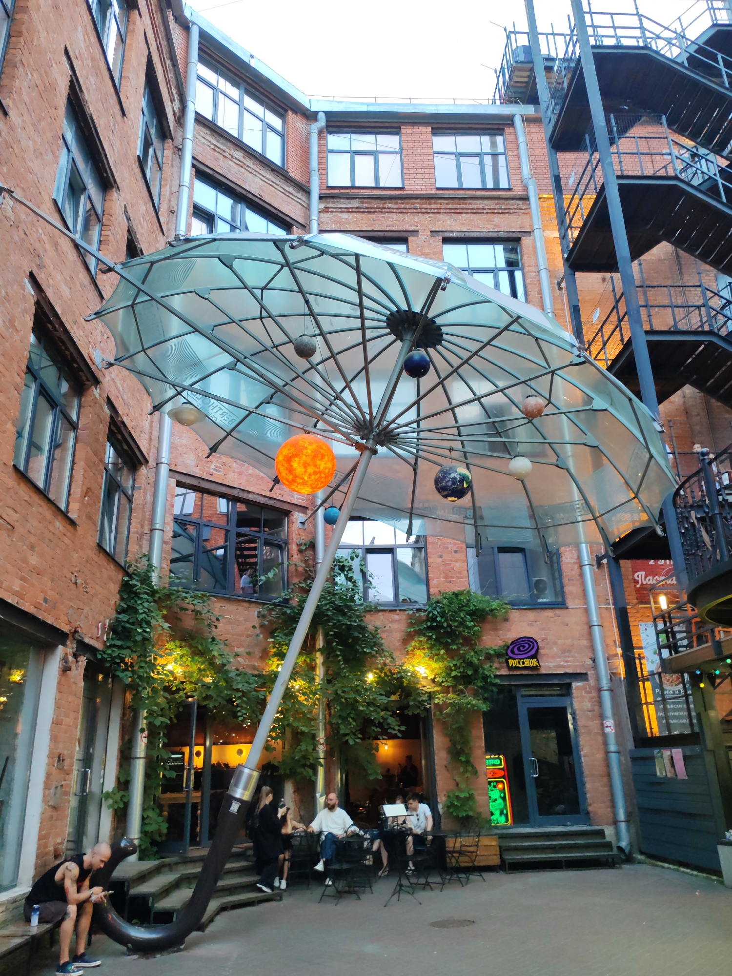 This is an outdoor courtyard scene in Saint Petersburg, Russia. The courtyard is enclosed by brick buildings with numerous windows. A large, translucent umbrella-like structure hangs in the center, adorned with several spherical objects resembling planets and a bright orange sphere that appears to be the sun. An ornate, dark metal staircase with multiple landings is attached to one of the buildings on the right. At the bottom of the staircase, a sign with Cyrillic text is visible. In the foreground, several people are seated at tables, seemingly at an outdoor cafe or restaurant. One man is sitting on a bench on the left, looking at his phone. Lush green vines with yellow lights are draped along the walls and entranceways of the buildings, creating an atmosphere of an urban garden. The lighting suggests it is either late afternoon or early evening.