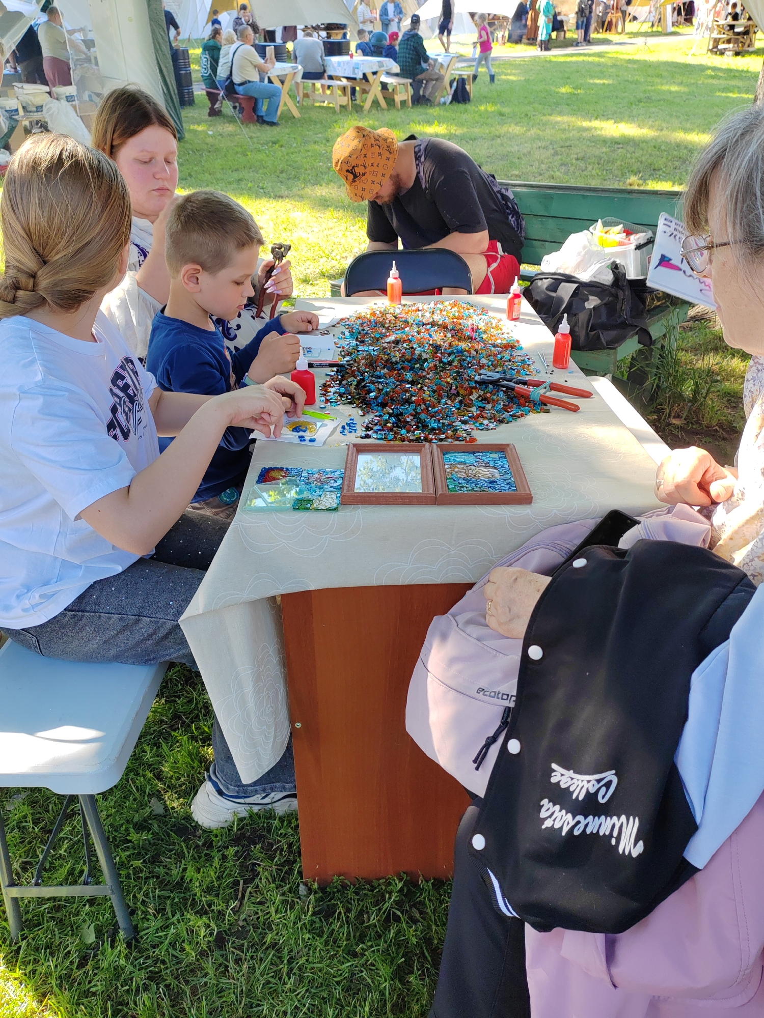 This image depicts an outdoor scene, likely at a festival or fair, with several people engaged in a craft activity. In the foreground, a table is set up with a large pile of colorful glass pieces, seemingly for mosaic art. Three individuals are visible around the table: a young boy in a blue shirt is focused on his work, a young girl in a white t-shirt is also participating, and an adult woman with blonde hair is partially visible. To the right, another adult, wearing glasses and a patterned scarf, appears to be observing or assisting. A stack of black and blue outerwear with "Minnesota College" embroidered on it is draped over a shoulder in the foreground. In the background, tents and other people are visible, suggesting a public gathering. The ground is covered in green grass.