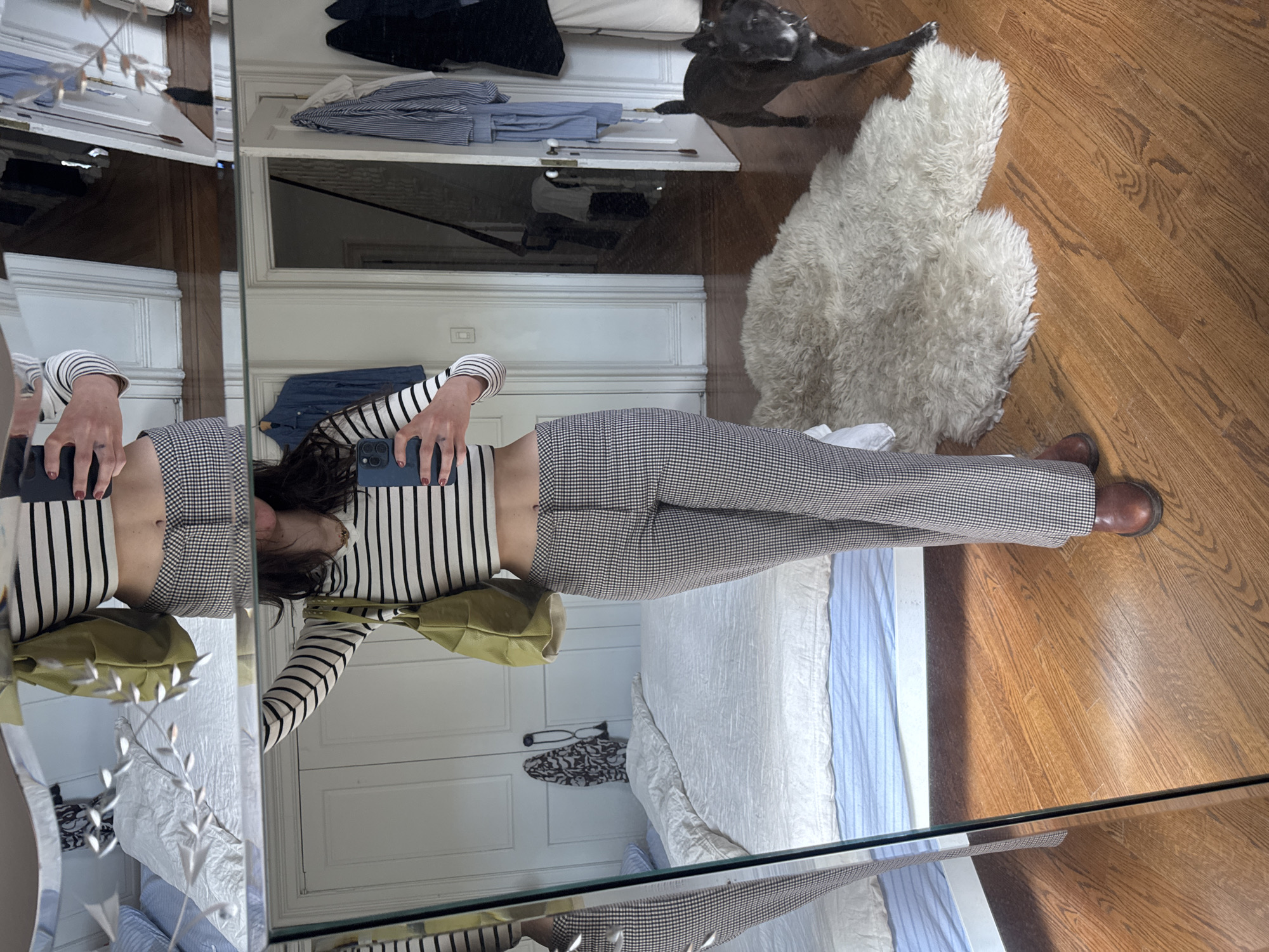 An indoor scene, likely a home in New York, United States, is captured through a full-body mirror selfie. The image features a person, presumably female, standing in front of a mirror, holding a dark-colored smartphone to take the picture. Her face is obscured by the phone. She is wearing a black and white striped long-sleeve crop top and fitted black and white checkered pants, paired with brown leather shoes. A light green shoulder bag hangs on her left side, and her nails are painted a dark color.
To the right of the person's reflection, a dark-colored dog, possibly a French Bulldog, is visible on the polished wooden floor, looking towards the camera. A shaggy white sheepskin rug lies on the floor nearby. The background reveals a domestic setting with a white bed featuring white and light blue striped bedding. An open white wardrobe or cabinet holds folded clothes, including light blue striped shirts and dark blue fabric. The room is brightly lit, indicating it is daytime. No discernible text is visible within the image.
