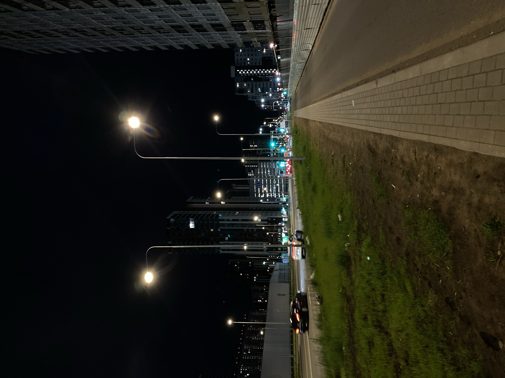 This is a nighttime photograph taken in Saint Petersburg, Russia, from a ground-level perspective looking down a road. The road has a paved sidewalk and a grass verge. The left side of the image is dominated by dark buildings and the glow of streetlights. Tall buildings, some with illuminated windows, can be seen in the distance. Cars with their headlights on are traveling on the road. The environment is urban, with a mix of built structures and greenery.