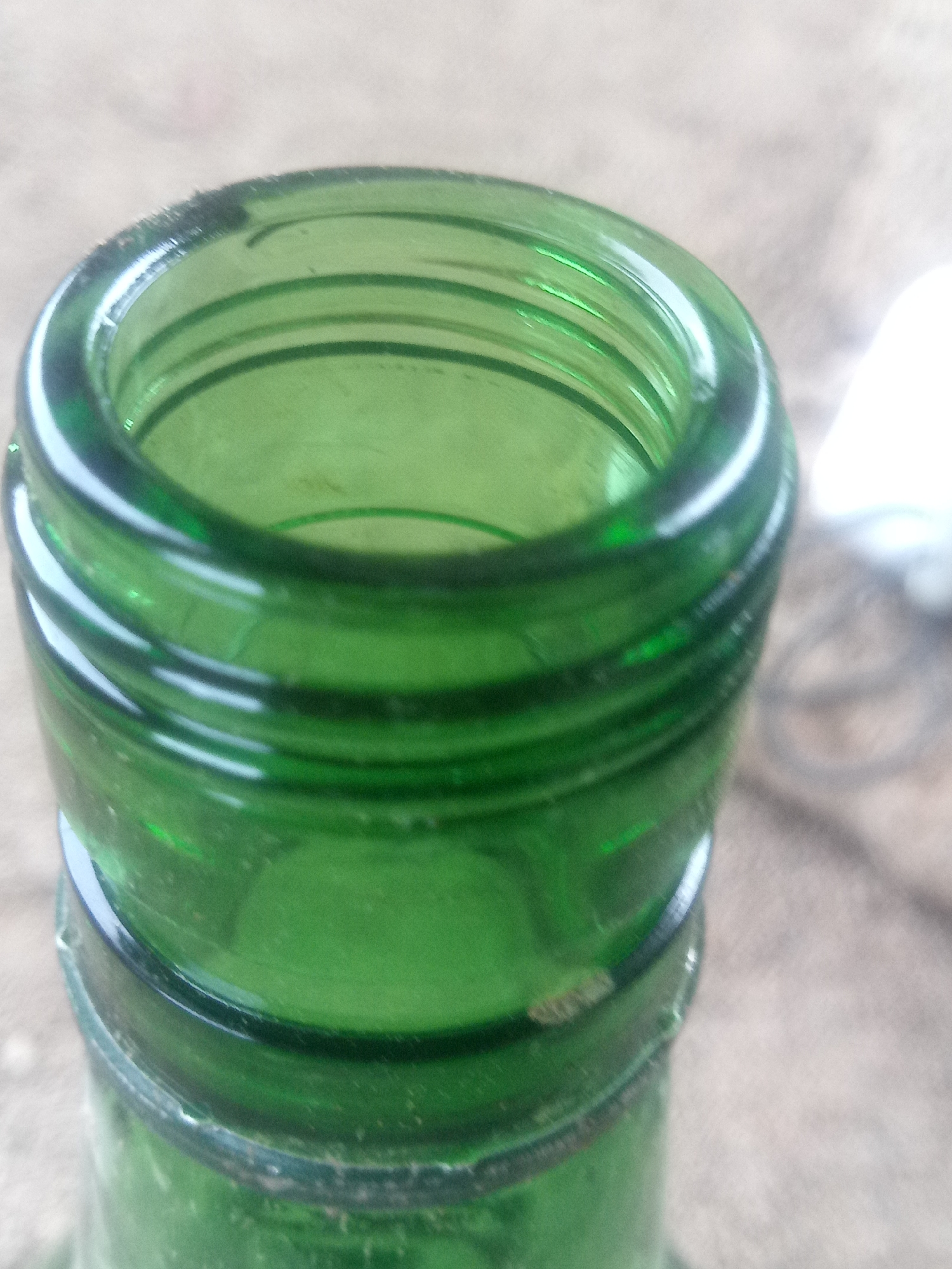 A close-up, eye-level shot focuses on the threaded opening of a green glass bottle. The bottle's opening is circular with a thick rim, and concentric ridges are visible on the interior. The glass appears to be slightly dusty. The background is out of focus and shows a textured, light-colored surface, possibly concrete or pavement, with some darker, indistinct objects. The lighting is natural, creating subtle reflections on the glass. There are no people or discernible actions visible. The location is not specified in the image.