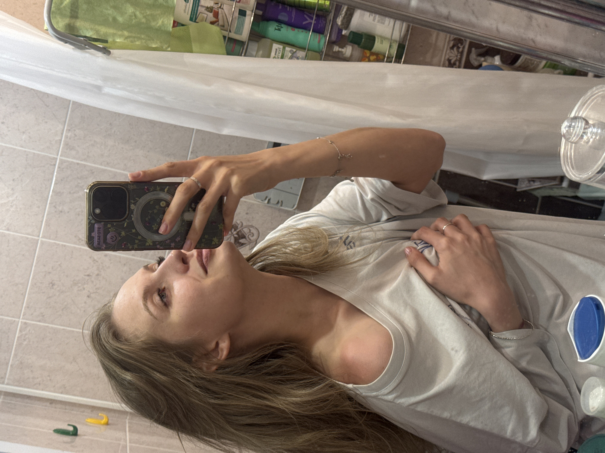 A woman is taking a selfie in a bathroom. She is holding a phone with a decorative case. The bathroom has tiled walls and a shower curtain. Various toiletries are visible in a wire rack. The location is potentially Izhevsk, Russia.