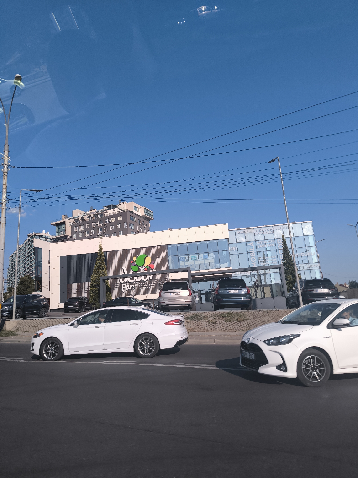 In Chișinău, Moldova, on a clear, sunny day, a street scene unfolds with a prominent modern building complex and various vehicles. The main building features a large, multi-story structure with a light-colored facade and large glass windows, possibly a commercial or residential complex. In front of this, a lower section of the building has a striking ribbed facade and a large, colorful logo that appears to be a stylized fruit or plant. The building's name, "Vipcar," is visible beneath the logo.
Several cars are present. A white Ford Fusion is in the foreground, traveling on the right side of the road, and a white Toyota Yaris is to its right, also in motion. Other vehicles, including a dark SUV, a dark sedan, and a silver SUV, are parked or moving in the background, suggesting a commercial area or parking lot. The sky is a vibrant blue, with a few wispy clouds and several power lines crisscrossing the upper portion of the frame. A street lamp stands on the right side of the image. The overall impression is a busy urban environment captured on a bright, clear day.