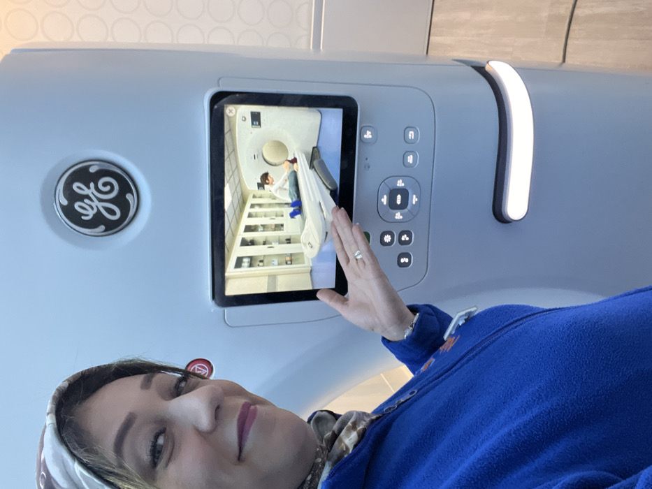 A woman takes a selfie in front of a medical imaging device, possibly a CT scanner. The device has a GE logo on its left side and a screen showing a scene of a medical professional assisting a patient in a similar imaging room. The woman is wearing a blue fleece jacket and a patterned headscarf. Her hand, adorned with a ring, is near the screen of the imaging device. The background is neutral and modern.