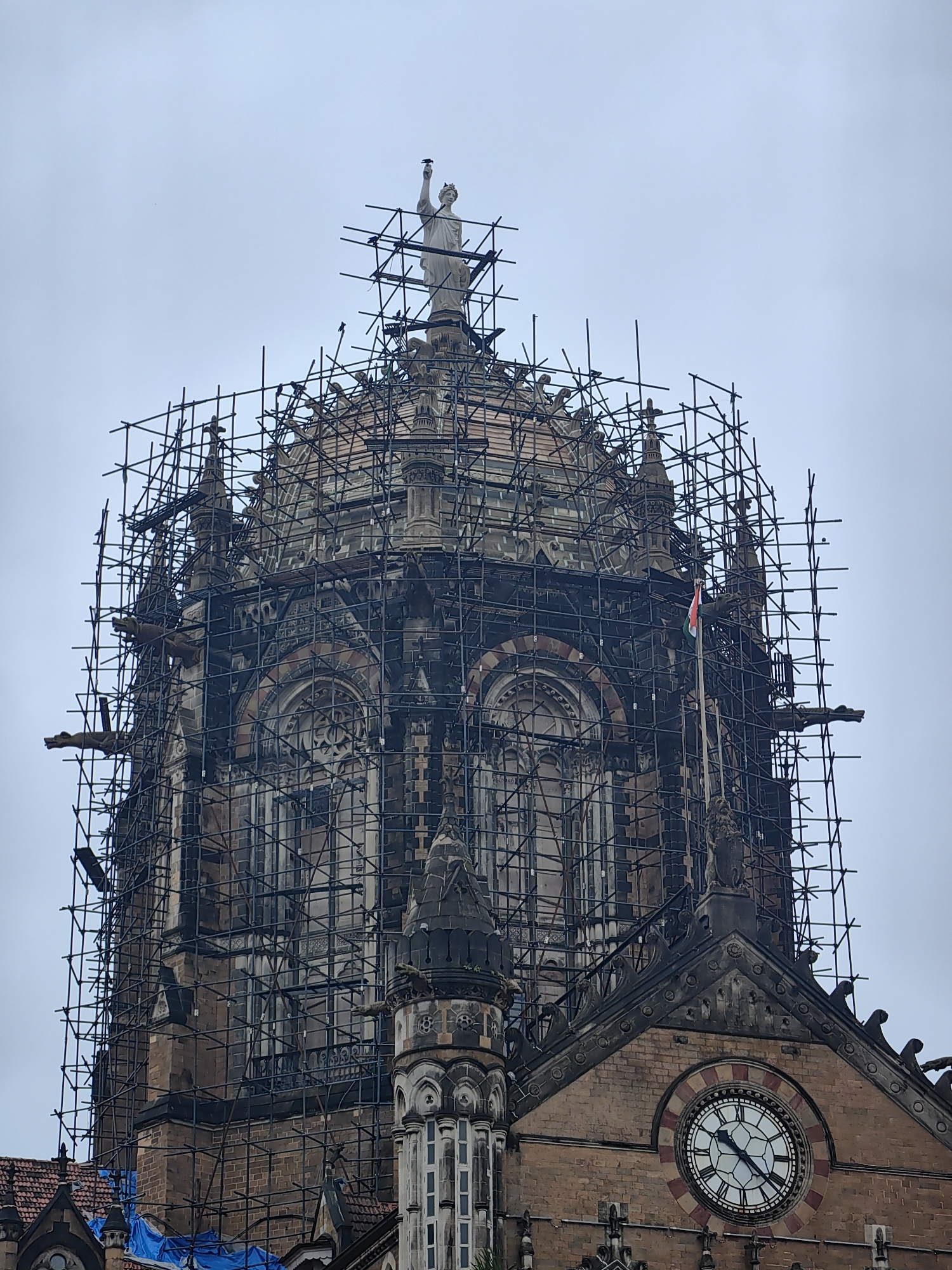 The image shows the Victoria Terminus railway station in Mumbai, India, which is also known as Chhatrapati Shivaji Maharaj Terminus. The building is covered in extensive scaffolding, indicating it is undergoing restoration or construction. A statue stands at the very top of the dome, with several birds perched on the scaffolding around it. An Indian flag is visible on a flagpole attached to the side of the building, partially obscured by the scaffolding. The building's architecture is detailed, featuring ornate stonework, arched windows, and gargoyles. A large clock face is visible on the lower part of the building. The sky is overcast, suggesting a cloudy day.