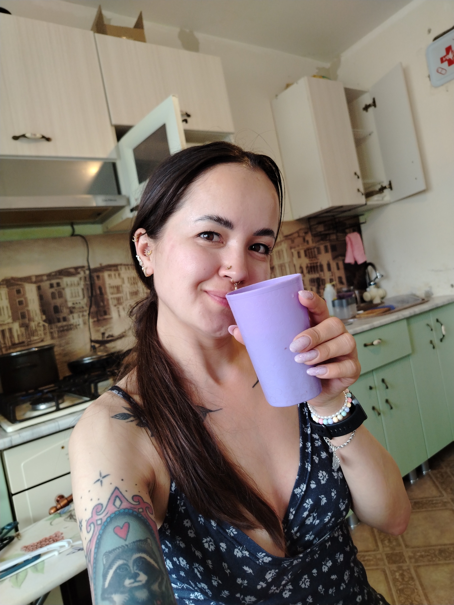 A selfie-style image shows a woman holding a purple cup. She is wearing a floral patterned top. She has a nose ring and several earrings. A tattoo is visible on her left arm. The background shows a kitchen with white and green cabinets, a stove, and a picture of Venice. The first aid kit on the wall indicates it is most likely located in Noviy, Russia.