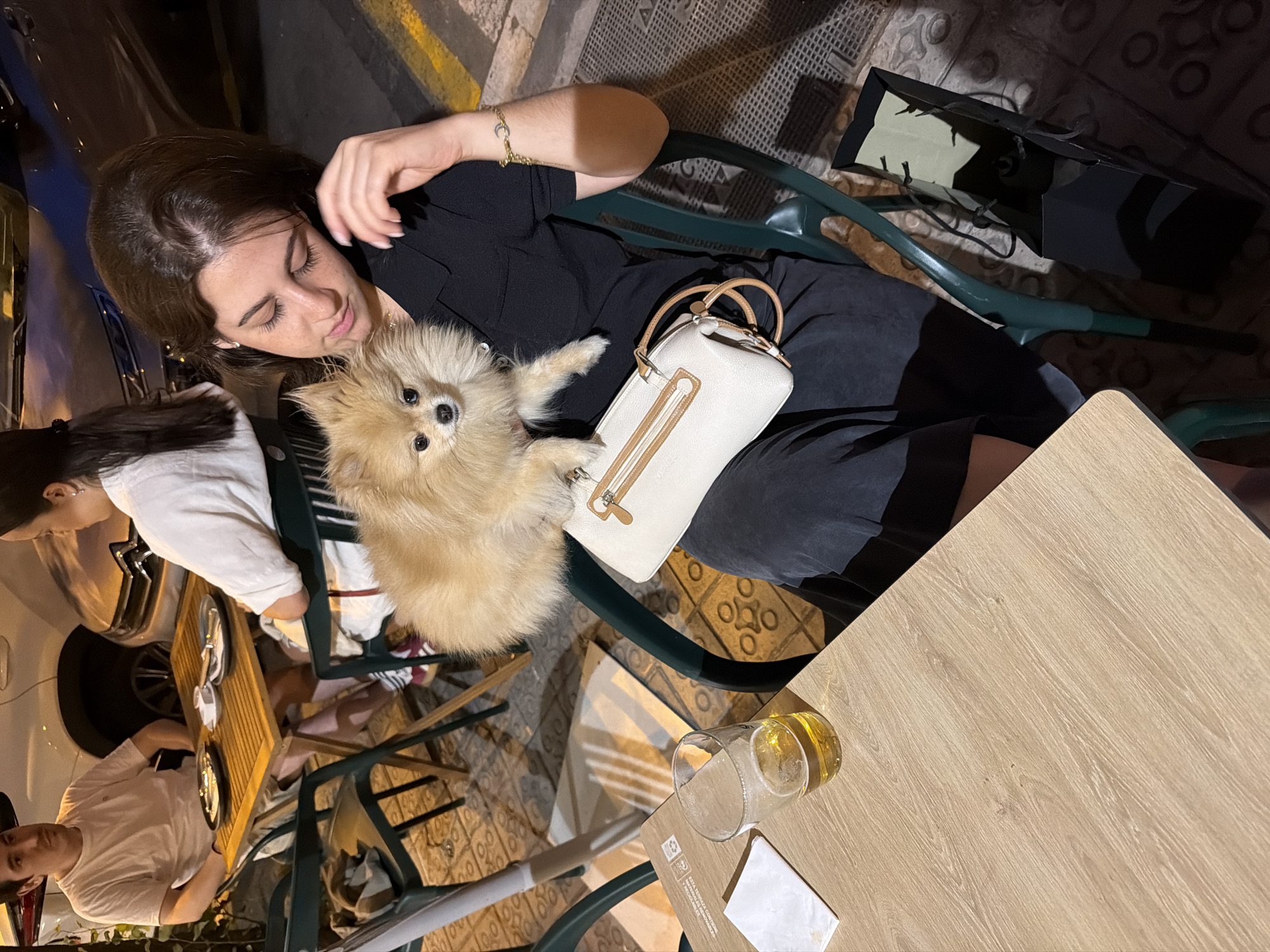 A woman sits outdoors at a table with a small dog in her lap. She is wearing a black collared shirt and a dark skirt. A white handbag with tan accents rests on her lap. A glass of beer and a napkin are on the wooden table in front of her. Behind her, a man in a white t-shirt sits at the same table. In the background, two people are seated at another table, and a white car is parked. The ground is paved with patterned tiles. The scene appears to be an outdoor cafe or restaurant in Barcelona, Spain.