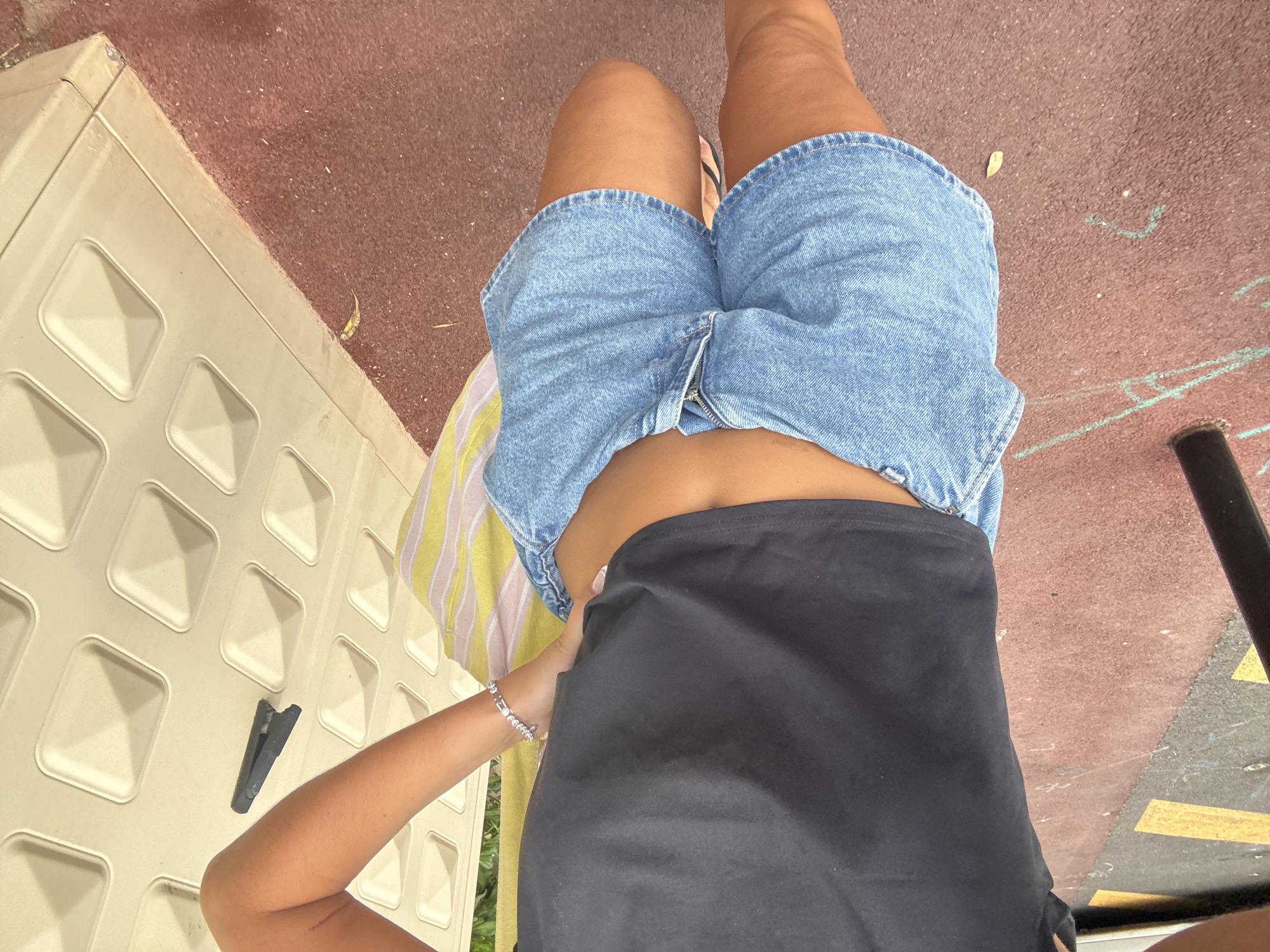 A person stands outdoors, dressed in a black crop top and light blue denim shorts. The person is holding a striped bag over their right shoulder and wearing a silver bracelet on their left wrist. They are standing on a reddish-brown surface, possibly a sports court or paved area, with yellow markings visible on the ground to the left. To the right, a beige wall with a textured pattern is visible, and a black clip is attached to it. The image appears to be taken in the city of Cannes, France.
