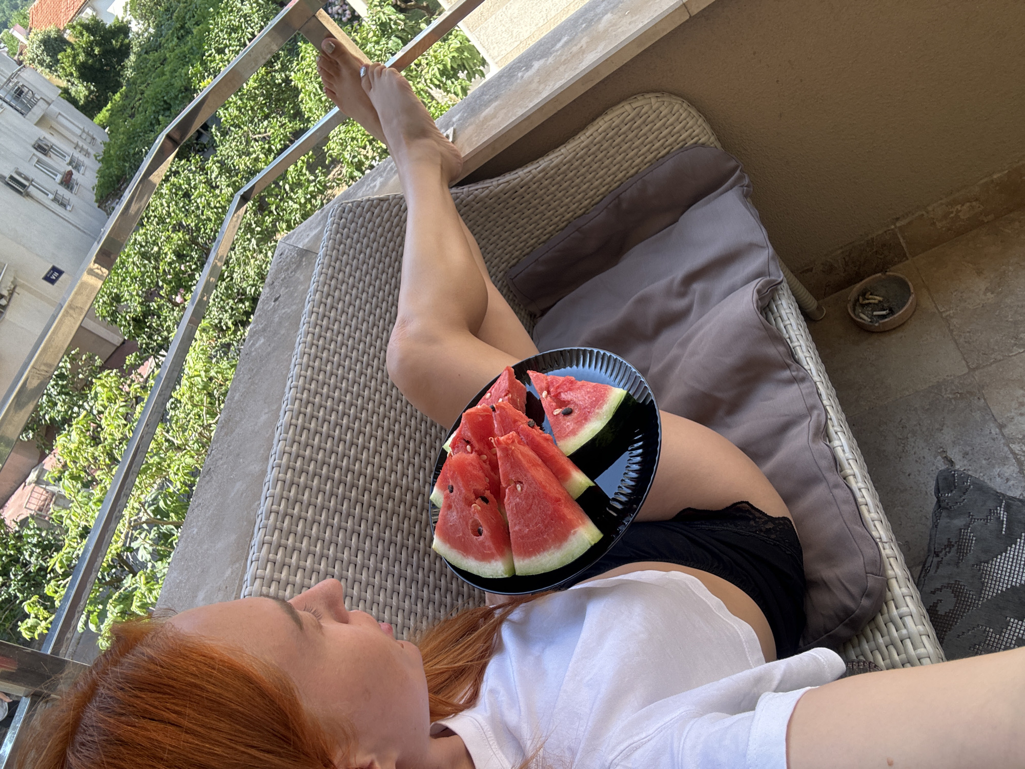 A woman with red hair is relaxing on a wicker chair on a balcony, holding a plate of watermelon. She is wearing a white t-shirt and black shorts. Her legs are crossed and extended, with one foot resting on the balcony railing. The scene is bathed in sunlight, suggesting it is daytime. In the background, there is a lush green landscape with trees and buildings. The railing of the balcony is made of metal. An ashtray filled with cigarette butts is visible on the floor next to the chair. The overall impression is one of leisure and relaxation in a warm, sunny environment.