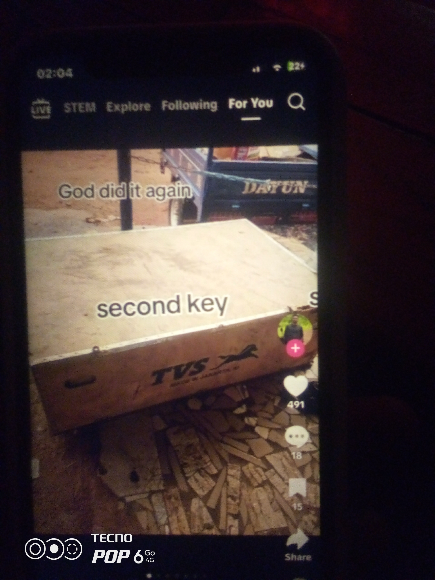 The image is a screenshot of a mobile phone displaying a video. The video shows a light brown wooden surface, possibly a box or lid, with the words "God did it again" and "second key" written in white text. A logo with a leaping animal, possibly a horse or panther, and the text "TVS MADE IN JAPAN" is visible on the surface. A partial view of a truck with the word "DAYUN" on it is also visible in the background. The phone screen shows a timer "02:04", navigation tabs "LIVE", "STEM", "Explore", "Following", "For You", and a search icon. There are social media interaction icons on the right side of the screen: a heart with "491", speech bubbles with "18", a bookmark with "15", and a share icon. The bottom of the phone displays "TECNO POP 6 4G". The image is dimly lit, suggesting it was taken in low light conditions, and the phone is being held, indicated by the slight angle. The city of Howayi, Nigeria is not depicted in this image.