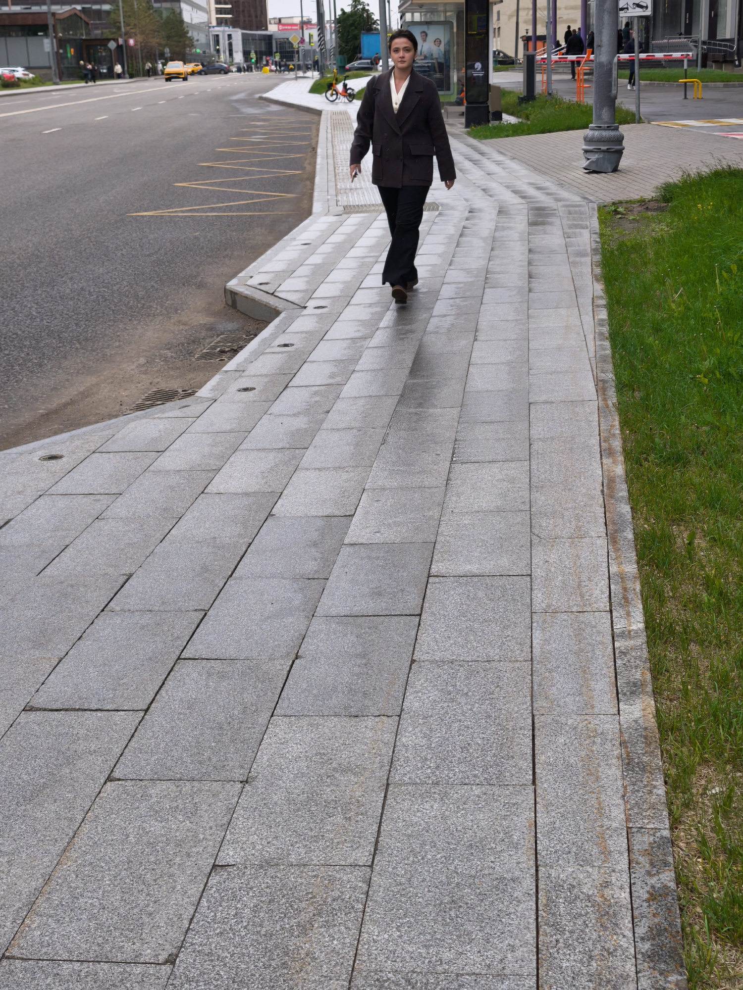 A woman in a dark suit walks towards the viewer along a wet, gray-tiled sidewalk in Moscow, Russia. The sidewalk is bordered by a street on the left and a patch of green grass on the right. The street is lined with buildings, and there is a yellow taxi visible in the distance. A bus stop is present on the sidewalk behind the woman. A barred traffic barrier stands in the far right background. The scene is overcast.