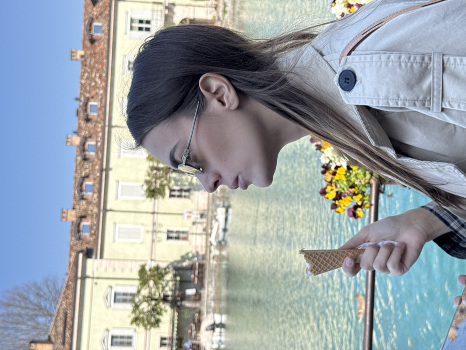 An outdoor scene in Peschiera del Garda, Italy, captures a female subject in profile, looking right, with a canal and historic buildings in the background. The female, centered in the frame, possesses long, dark brown hair and wears round, silver-framed Miu Miu sunglasses with light lenses. She is dressed in a light-beige trench coat over a partially visible plaid shirt. In her left hand, in the lower foreground, she holds a partially eaten waffle ice cream cone containing white ice cream.
The background features a body of turquoise-colored water, likely a canal, extending horizontally across the mid-ground. Multiple small, light-colored boats are moored or floating on the water's surface. A single brown duck is visible floating on the water in the lower-mid-left. On the far side of the water, a large, historic building with light yellow walls, a red-tiled roof, multiple windows with white shutters, and several chimney pots is prominent. Behind the building, bare deciduous trees are visible against a clear blue sky. In the lower right, a blurred planter box contains yellow, red, and purple pansies. The scene is illuminated by natural sunlight.
