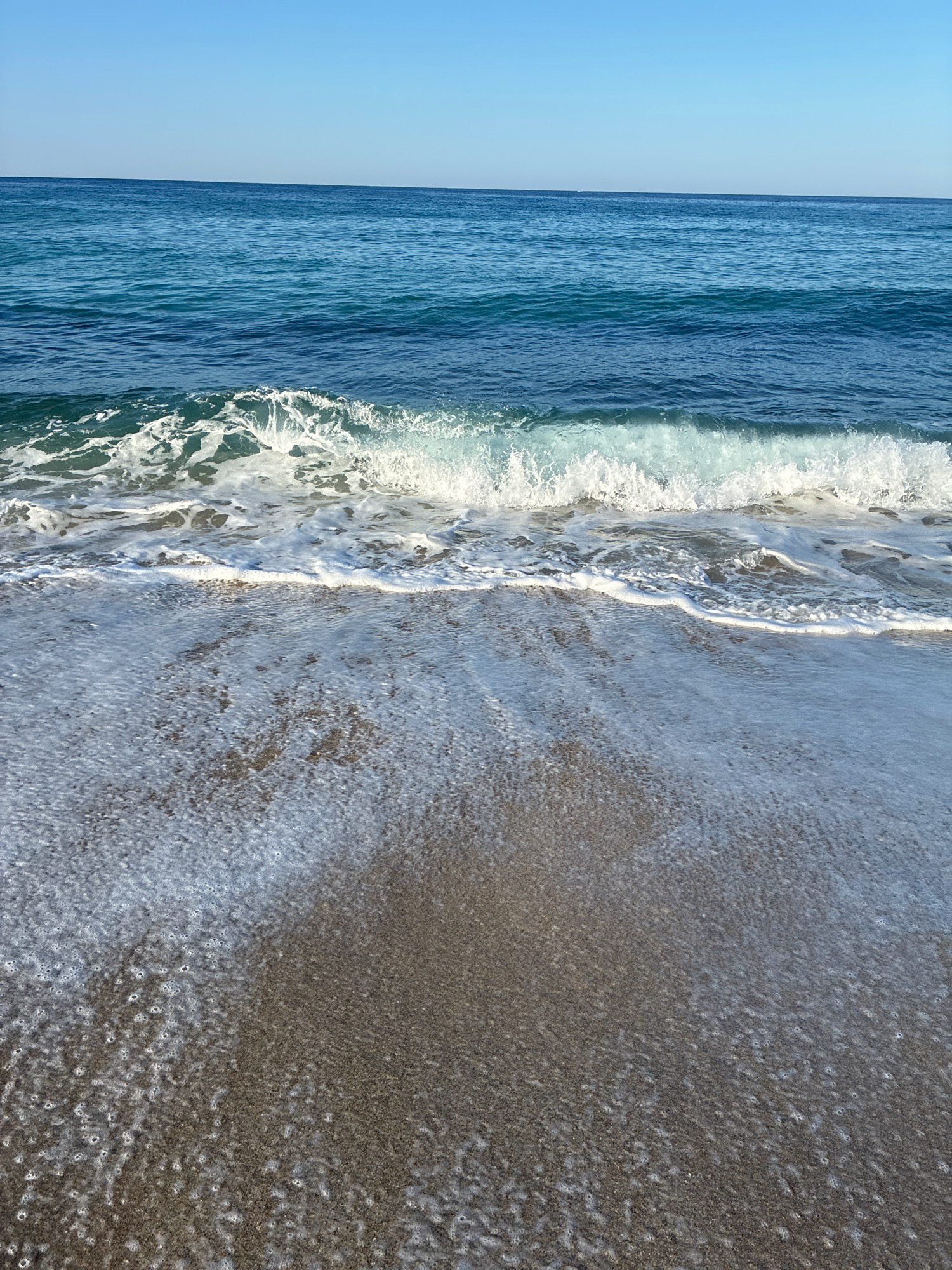 A tranquil ocean scene unfolds with a clear, bright blue sky above a calm sea that transitions from a deep sapphire to a lighter turquoise. A gentle wave crests and breaks, producing white foam that washes onto a sandy shore. The wet sand at the water's edge is textured with the imprint of the receding foam. The image captures a serene moment at a beach in Alanya, Turkey, characterized by its natural beauty and peaceful atmosphere.