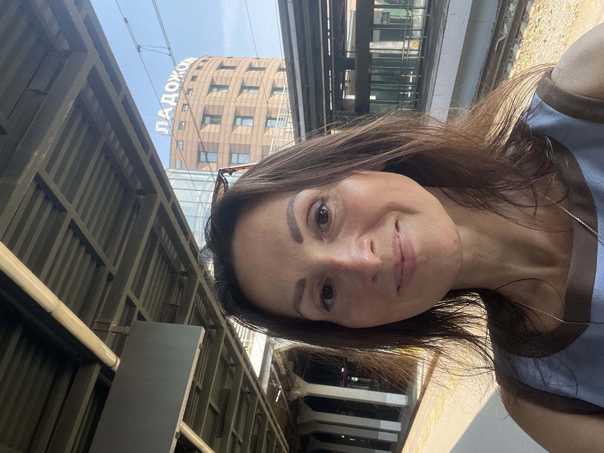 A woman with brown hair and brown eyes, wearing a blue and brown top, takes a selfie at a train station. Behind her, a curved, multi-story building with windows is visible, featuring a sign with Russian lettering that reads "ЛАДОЖСКИЙ". Overhead, a train station roof structure with metal beams and a light-colored pipe is present. The environment appears to be outdoors on a sunny day. The location is Vsevolozhsk, Russia.