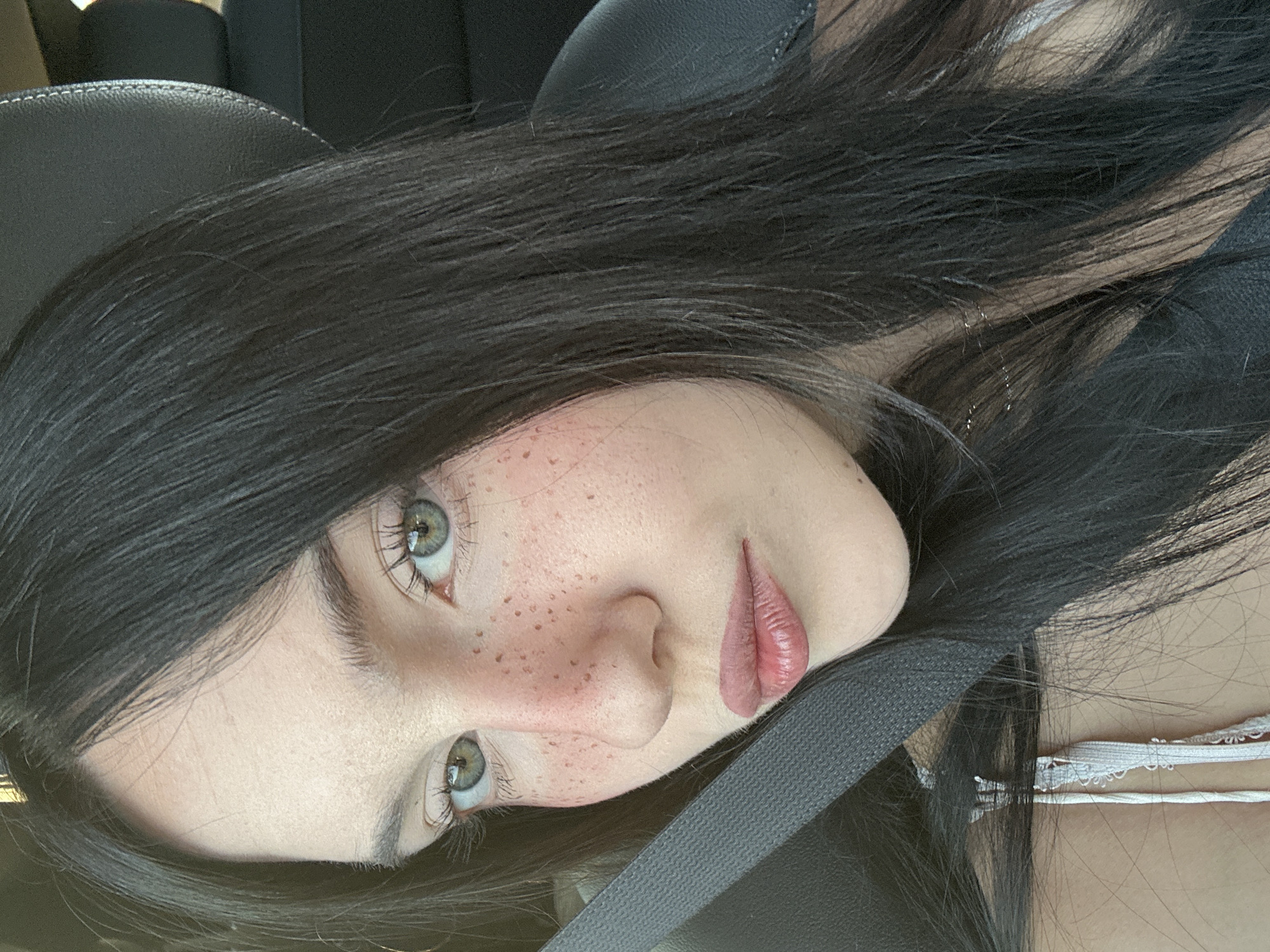 A young woman with long, dark hair and light green eyes is shown in a close-up, side-profile shot. She has a fair complexion with numerous freckles across her nose and cheeks. Her lips are slightly parted and have a glossy, pink lipstick applied. She is wearing a white lace-trimmed top, and a grey seatbelt is visible across her chest. The background appears to be the interior of a car, with a black leather headrest and seat visible. The lighting suggests it is daytime, possibly with sunlight filtering in. The image does not provide any specific visual cues or context that would allow for determination of its location within Grottammare, Italy.