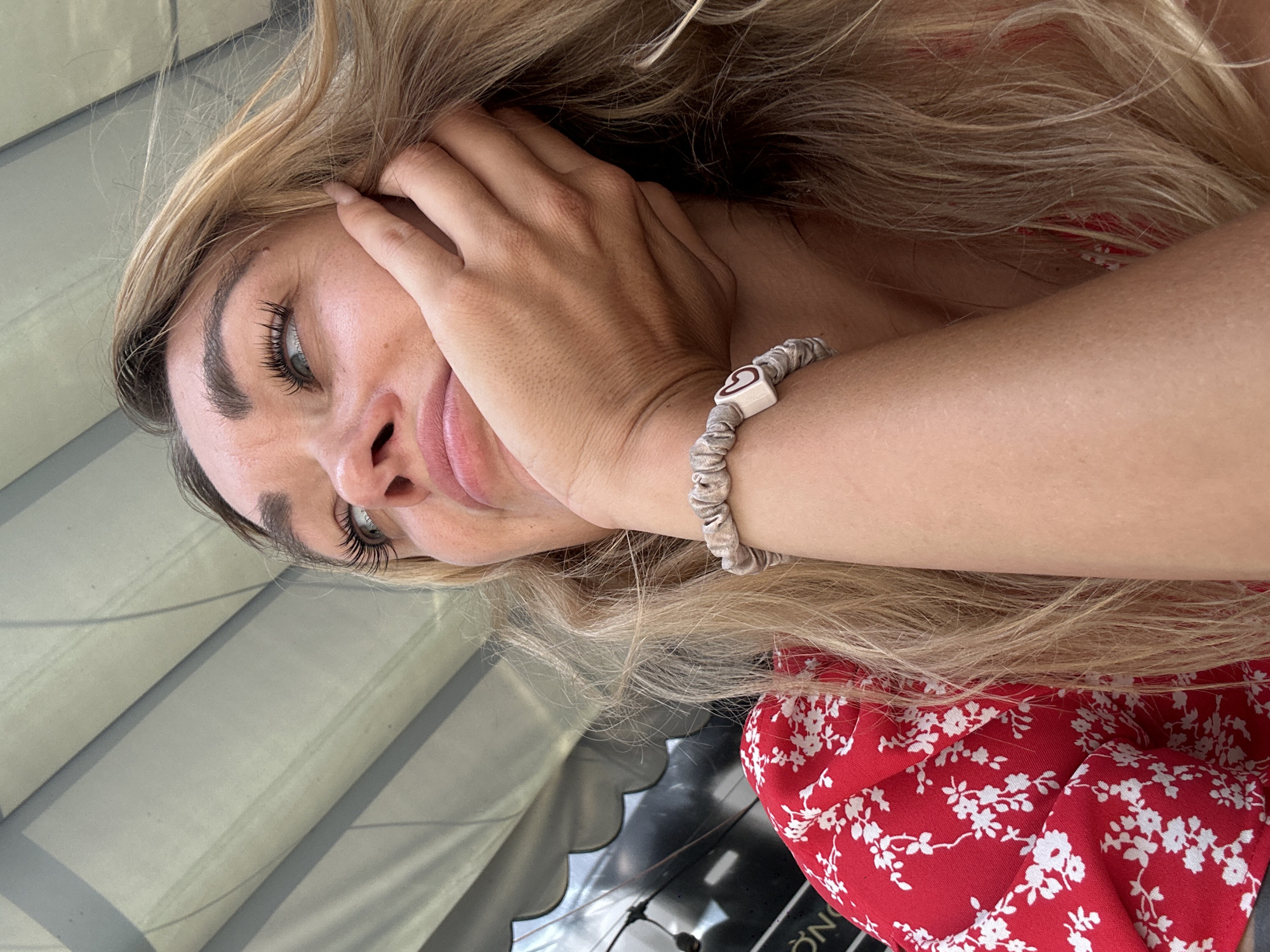 A young woman is depicted in a photograph, seemingly taken at a high angle. The woman has blonde hair that cascades around her face and onto her arm. She is wearing a red floral-patterned top. Her right hand is raised and gently touching her face near her temple. A bracelet with a possible heart-shaped embellishment is visible on her wrist. The background contains what appears to be a window shade and some other indiscernible objects. The location is unspecified in terms of a city or other geographic location, as no readily identifiable details are provided. No evidence indicates that this photo was taken in Đồng Nai, Vietnam. The description focuses solely on the visible elements in the image.