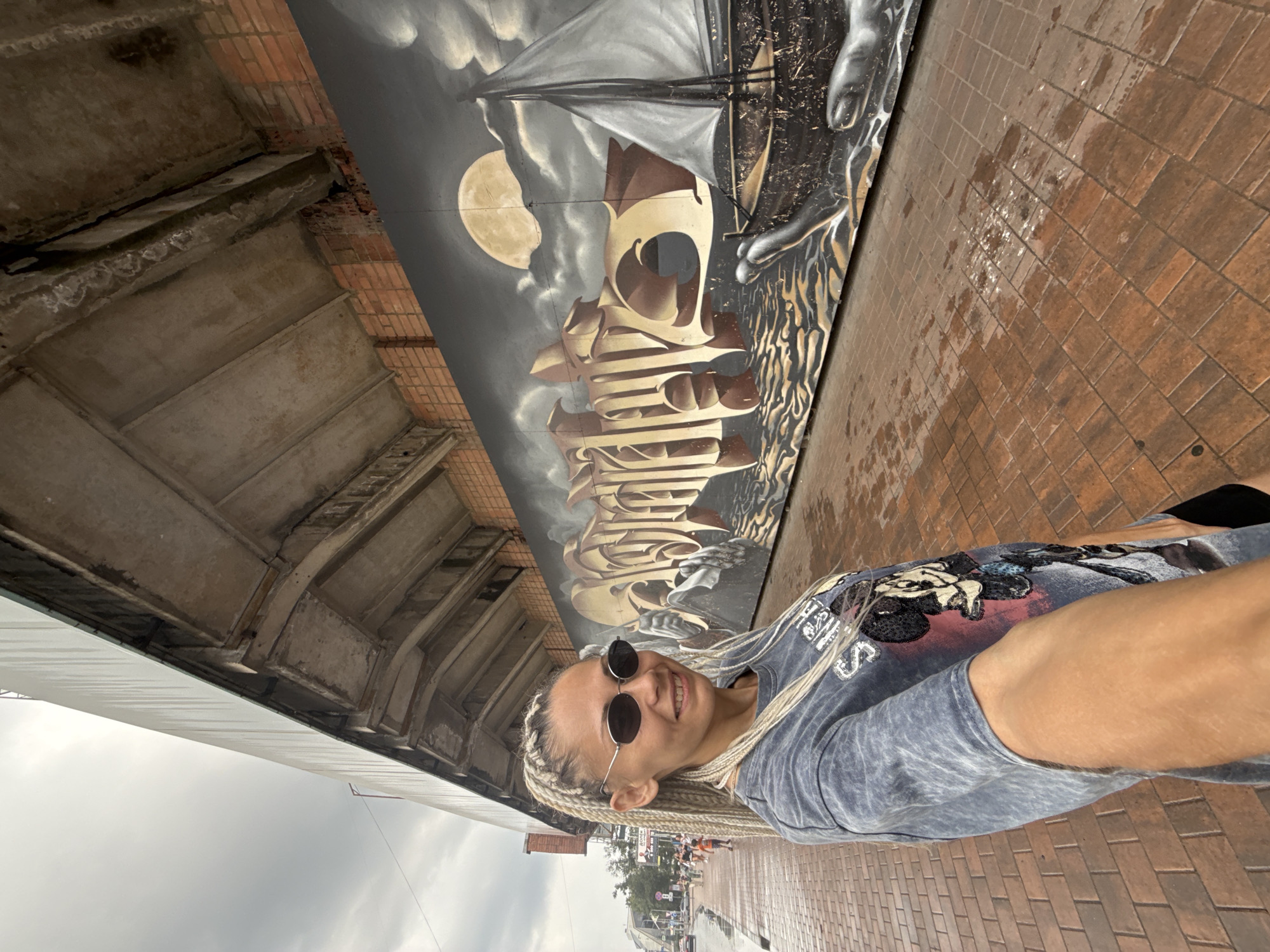 A woman with blonde braided hair and sunglasses smiles at the camera. She is wearing a blue t-shirt with a Mickey Mouse graphic. She is standing on a wet brick sidewalk next to a mural. The mural depicts a sailboat, a moon, and what appears to be a large hand. The scene appears to be outdoors. The sky is cloudy, suggesting overcast weather. The brick pathway is damp, indicating recent rain. In the background, buildings and trees are visible, as well as other people walking along the sidewalk. The overall impression is a casual, outdoor snapshot, possibly taken during the daytime. There is text on the woman's t-shirt, part of which is visible. The mural itself features stylized lettering and imagery. The setting suggests an urban or semi-urban environment.