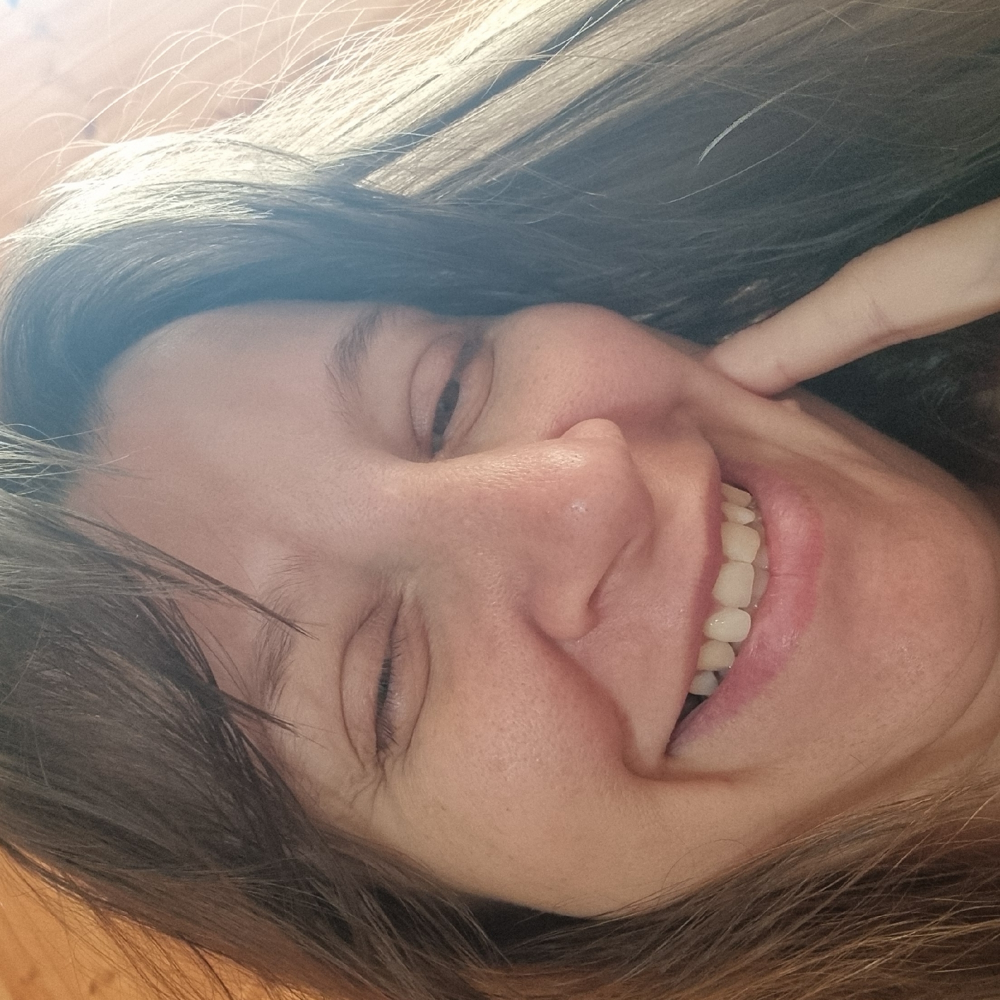 This is a close-up, head-and-shoulders selfie of a woman with brown hair and fair skin. She is looking directly at the camera with a playful expression, her left eye squinted and her right eye partially closed. Her mouth is open in a wide smile, revealing white teeth. Her right hand is touching her cheek. The lighting suggests an indoor setting, possibly near a window, with warm light illuminating her face. There is no discernible information about the location within Dunnae-myeon, South Korea.
