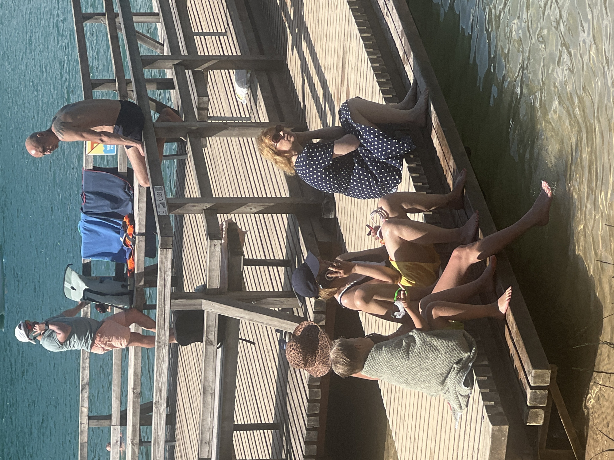 On a sunny day, several people are gathered on a wooden pier on the shore of a body of water. In the foreground, a child wrapped in a green blanket sits on the edge of the pier, dipping their feet into the water. Next to the child, an adult in yellow shorts and a blue hat is talking on a mobile phone. Further back and to the right, a woman in a dark blue polka dot dress is sitting on the pier, looking to her left. In the background, a man with tattoos is sitting on the railing of the pier, looking out at the water, and another man in a green t-shirt and pink shorts is standing and also talking on a mobile phone. There are towels and bags on a bench behind him. The pier is constructed of weathered wooden planks. The water is clear and shallow near the pier, with sandy bottom visible. The location is the bathing area of Lake Tapeliai in Vilnius, Lithuania, as indicated by a sign on the pier.