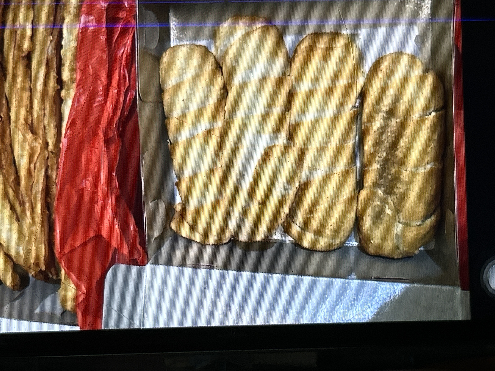 The image shows a box of baked goods, possibly pastries, arranged in two layers within a cardboard box. The top layer appears to be a stack of golden-brown, long, and somewhat flat items, partially obscured by a bright red plastic wrapper that is folded over them. The bottom layer contains at least four elongated, cylindrical pastries with a golden-brown, slightly uneven surface, exhibiting lighter streaks or patterns. These pastries are arranged side-by-side. The box is white with visible blue lining on the interior walls. The image is taken from a slightly elevated angle. No specific location in Depok, Indonesia, can be determined from the visual information provided.