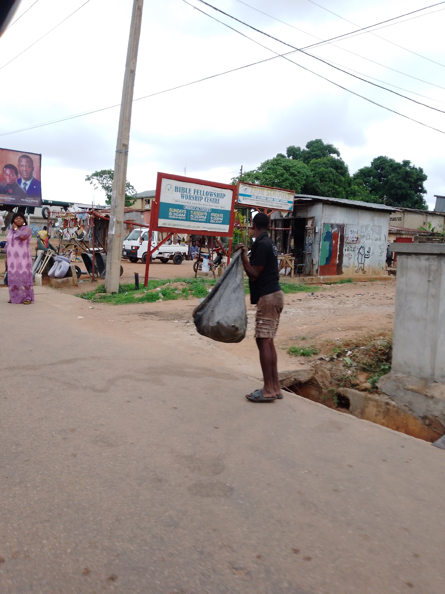 The image is taken in Sabon Tasha, Nigeria, and depicts a street scene. A man, dressed in a black shirt and khaki shorts, stands on the edge of a road holding a large, gray bag. Across the street, a woman in a pink dress walks along the roadside. In the background, there is a sign for "BIBLE FELLOWSHIP WORSHIP CENTRE." A small white truck is parked nearby, and a shed-like structure is visible with graffiti on the walls. The environment appears to be a somewhat urban or semi-rural setting.