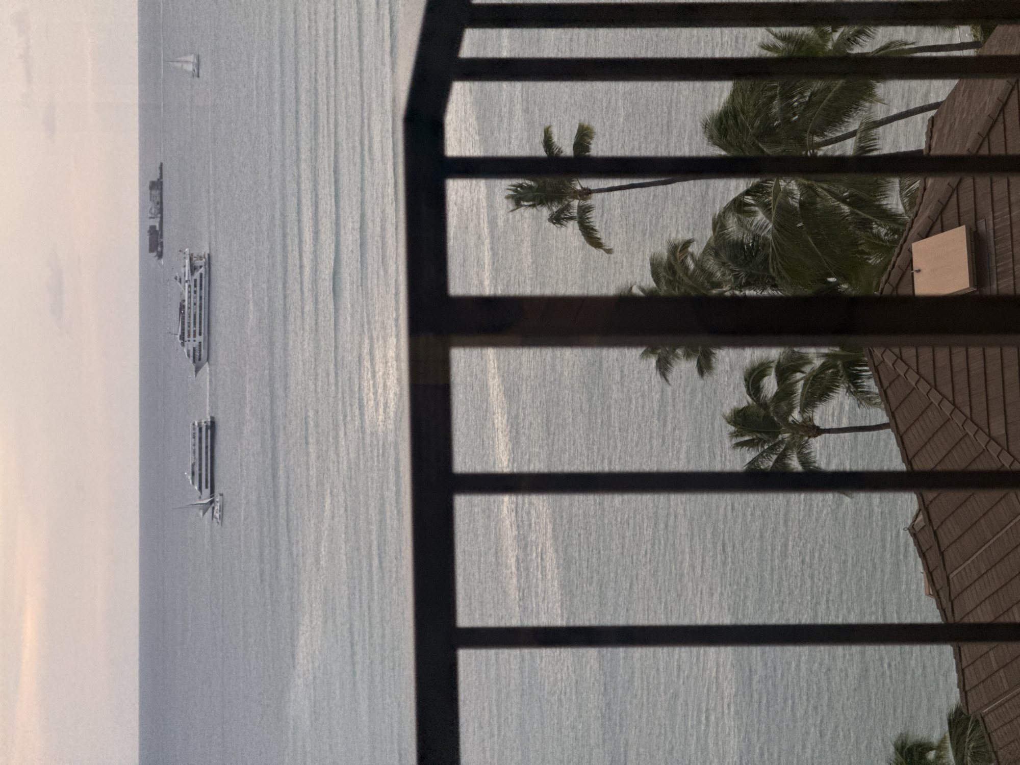 The image shows a view from what appears to be a balcony, likely in Honolulu, United States of America. The foreground is dominated by a dark, vertically-oriented railing. Through the railing, the scene unfolds: the ocean takes up a significant portion, with several ships and a sailboat visible on the left side of the frame. Additionally, palm trees are scattered, interspersed with a glimpse of rooftops in the vicinity.