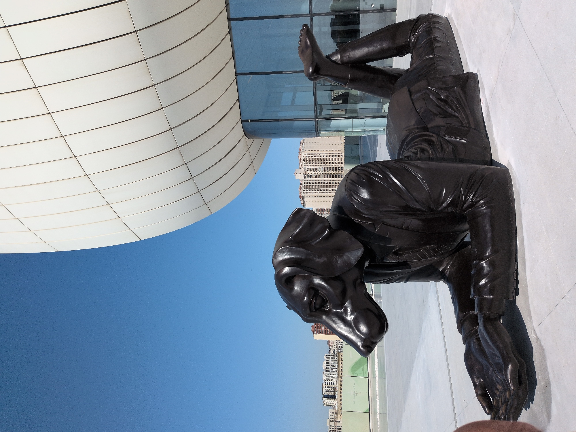 This is a full-body, low-angle, outdoor shot of a dark bronze sculpture of a dog wearing a suit, lying on its stomach with its legs crossed at the ankles and its arms bent at the elbows, with its hands open in front of it. The sculpture is on a white, tiled surface, and behind it is the exterior of a modern, white, curved building with large glass windows. In the background, there are several multistory buildings and a clear blue sky. The sculpture is a statue of a dog on its stomach. The sculpture is located in Baku City, Azerbaijan, in front of the Heydar Aliyev Center.