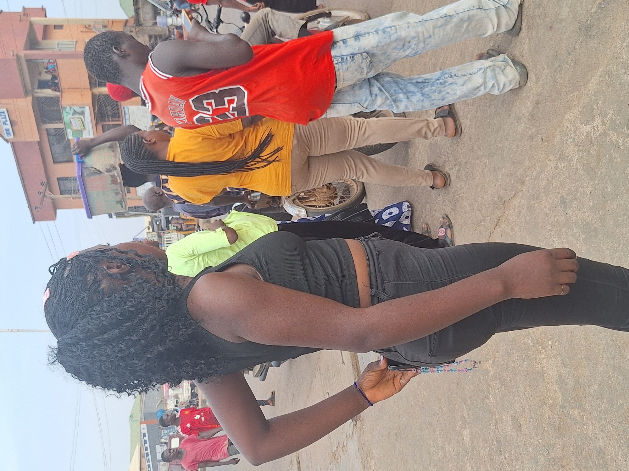 The image depicts an outdoor scene in Sabon Tasha, Nigeria, likely a street or public area. Several people are visible, including a woman in the foreground wearing a black top and jeans, holding a mobile phone. Behind her, there are other individuals wearing different colored shirts (red, yellow, and light green). One person wears a red jersey with the number 23. A motorcycle is also visible. The environment includes buildings and a somewhat overcast sky. The ground appears to be paved.