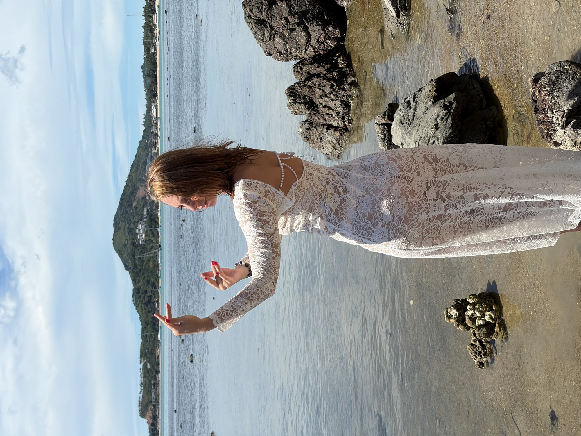 A woman stands on a rocky shore in Baan Bo Phut, Thailand, wearing a white lace dress. She is facing away from the camera, with her head turned to her left, looking out at the ocean. Her arms are raised, and her hands are positioned in a gesture. The water is calm, and there are boats visible in the distance. The sky is partly cloudy.