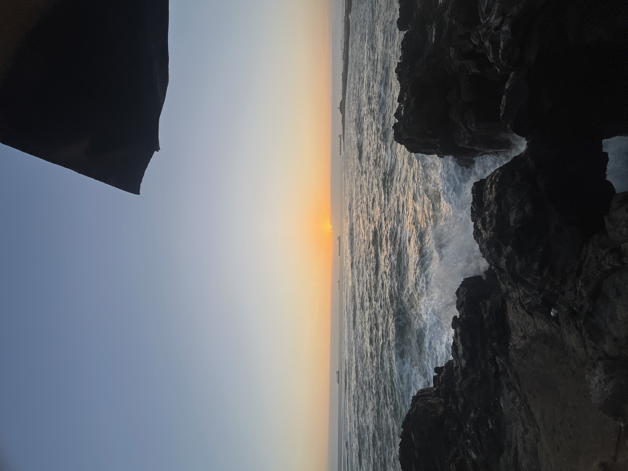 The image is an angled shot of a shoreline at dusk or dawn. The sky transitions from light blue to orange. The sea is choppy with white water where waves crash against dark, craggy rocks. In the distance, ships are visible on the horizon. A dark object, possibly part of a building or structure, is in the upper left corner. The scene likely captures the rocky coastal area of Arica, Chile, known for its beaches, cliffs, and dramatic sunsets over the Pacific Ocean.