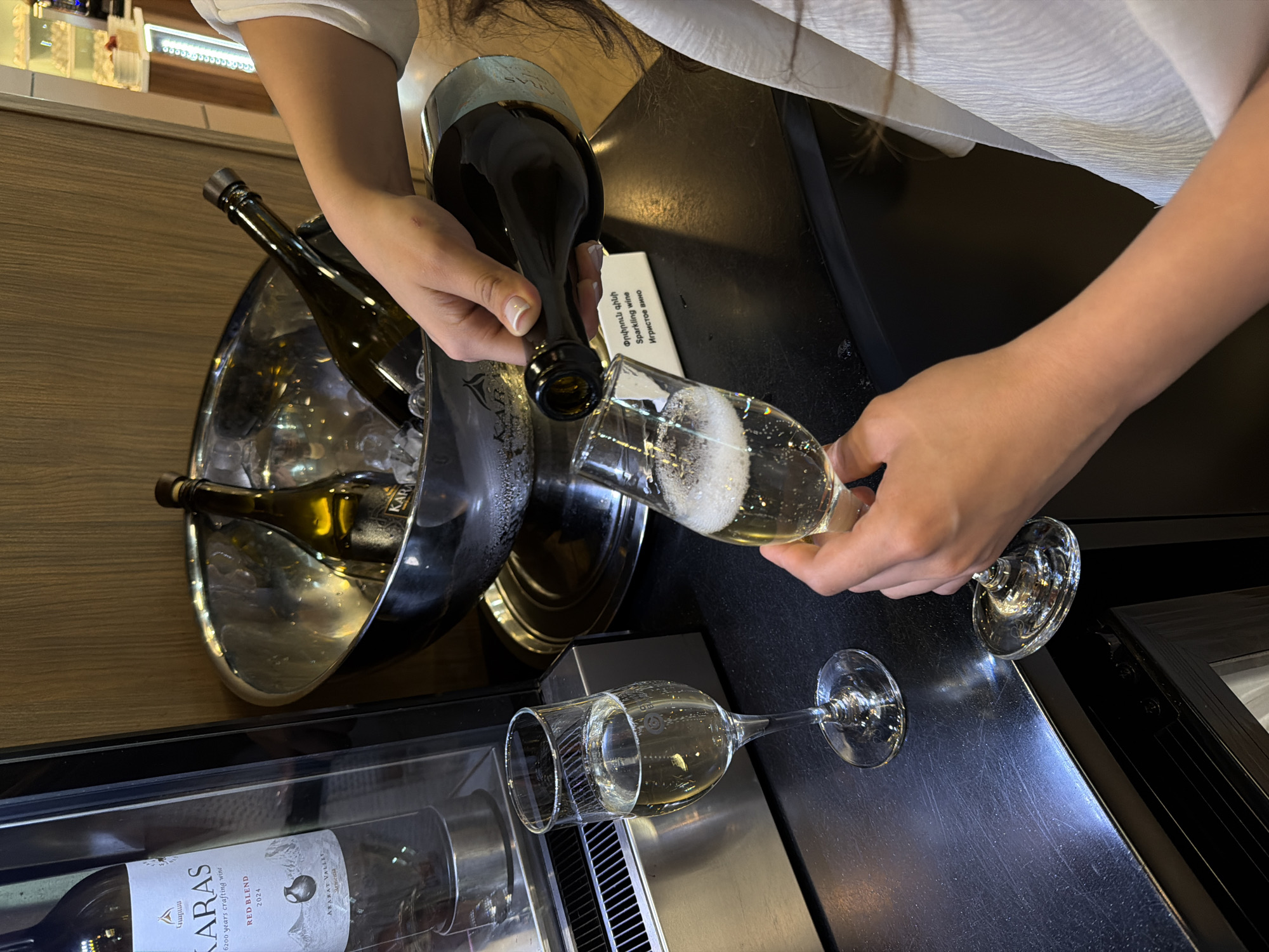 The image shows a person pouring sparkling wine into a glass. A silver ice bucket containing three bottles of wine stands on the left of the frame. Another partially filled wine glass lies on the surface in the foreground. A wine refrigerator is partially visible at the bottom. A card with the text "Sparkling wine" in three languages is placed near the glass. The wine brand is visible as "KARAS." The image seems to have been shot in Yerevan, Armenia, as the text on the wine label says "ARARAT VALLEY."