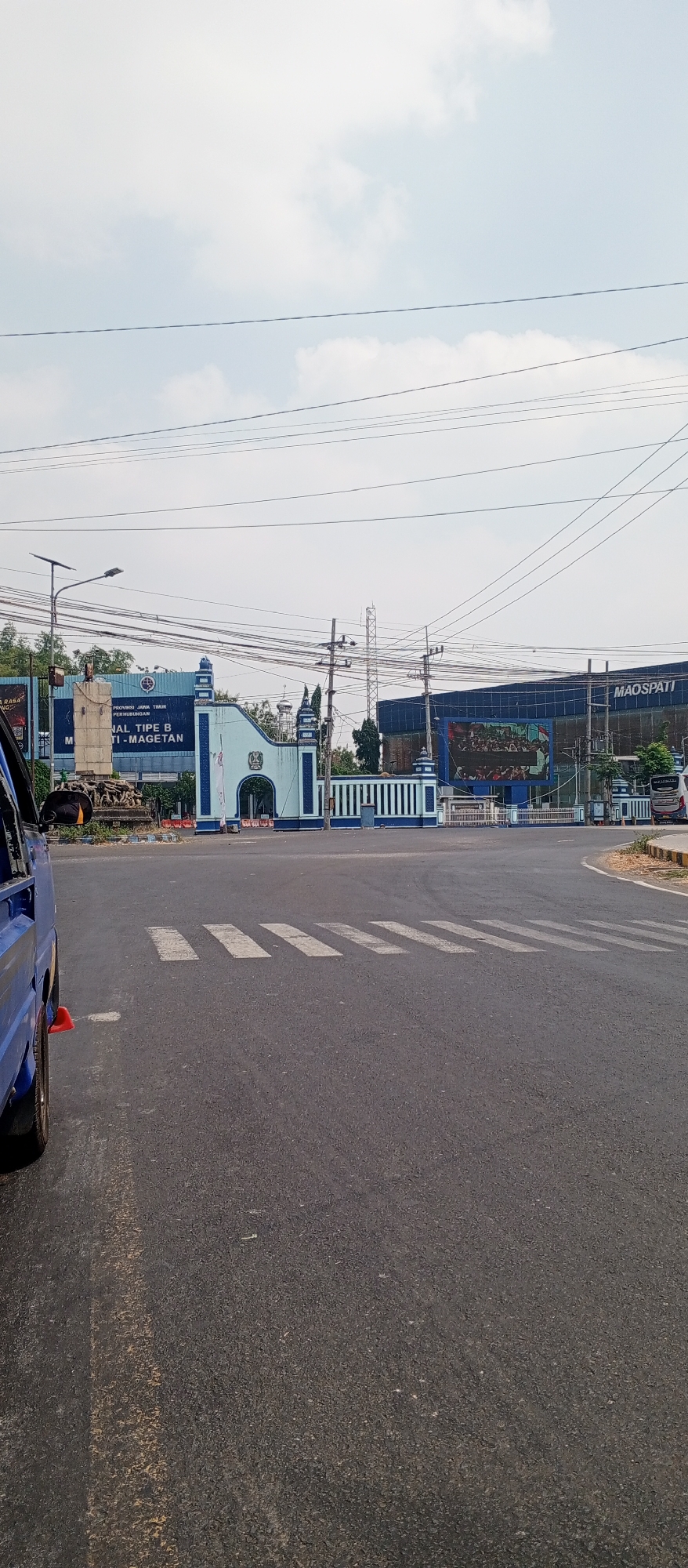 The image shows a view of a street in Maospati, Indonesia, on a cloudy day. In the foreground, the asphalt road is visible with white pedestrian crossing lines. A blue truck is parked on the left side of the road. In the middle ground, there is a blue and white gate with decorative elements and a sign that reads "PROVINSI JAWA TIMUR PERHUBUNGAN". To the right of the gate is a large building with a digital screen displaying a crowd of people. The building has the text "MAOSPATI" prominently displayed on its facade. The sky is overcast with visible power lines crisscrossing the frame.
