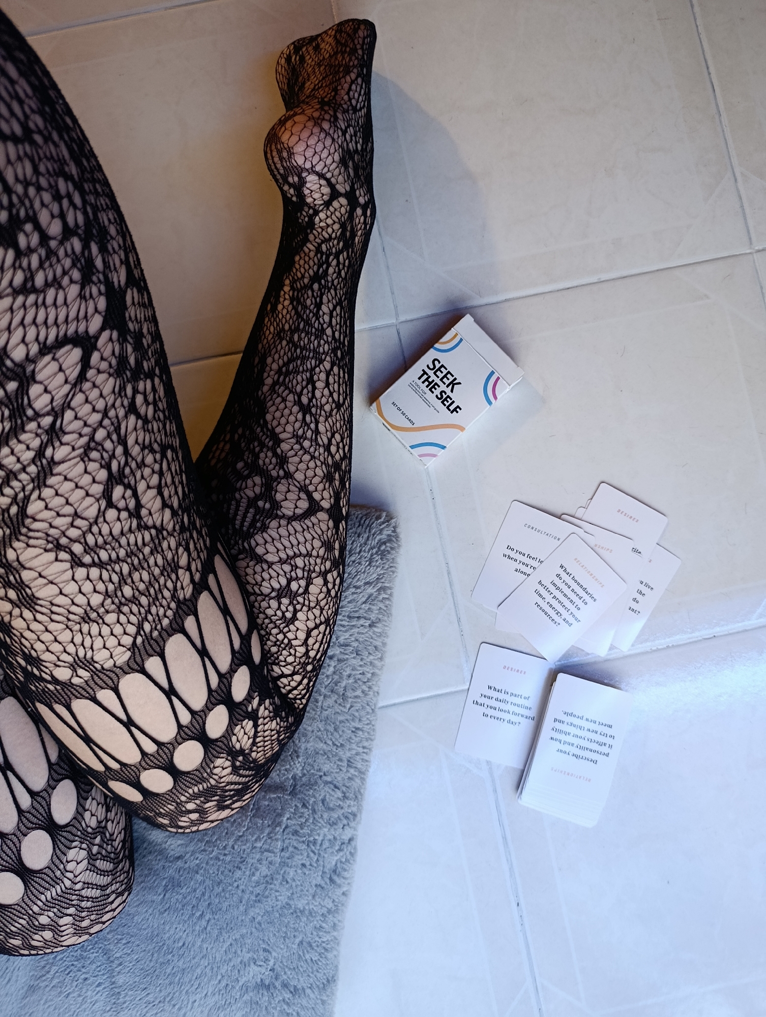 The image shows a pair of legs wearing black patterned tights resting on a gray fuzzy mat on a white tiled floor. Next to the legs are a deck of "Seek The Self" cards and several individual cards scattered around. The location is not specified and is unlikely to be Svilengrad, Bulgaria, without further context.