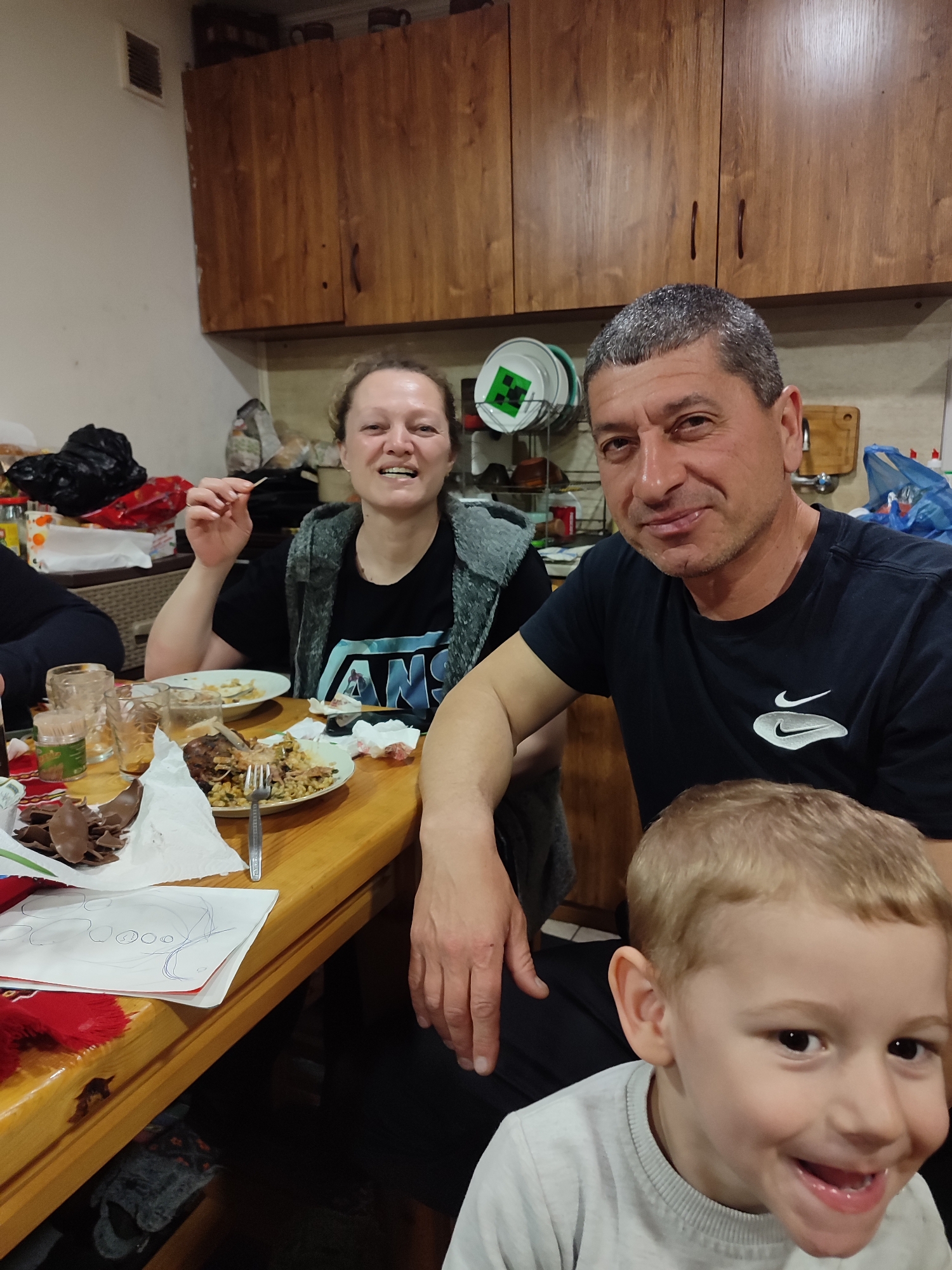 A family is gathered around a wooden table in a kitchen. The kitchen features dark brown wooden cabinets on the upper wall. On the table are plates with food, a fork, used glasses, a toothpick container, crumpled paper napkins, and pieces of chocolate. A piece of paper with a child's drawing is at the edge of the table.
A woman wearing a black Vans t-shirt and a grey jacket is sitting to the left of the frame, holding a toothpick. A man in a black Nike t-shirt is seated beside her, with his arm resting on the table. A young boy with blond hair and a grey sweater is positioned in the lower right corner of the image, smiling at the camera.
Plates with food are present, including meat and grains. Additional items like plates and some food items are on the wall behind the man. A green and white plate hangs on the wall.
The location is possibly a domestic setting in Varshilo, Bulgaria.