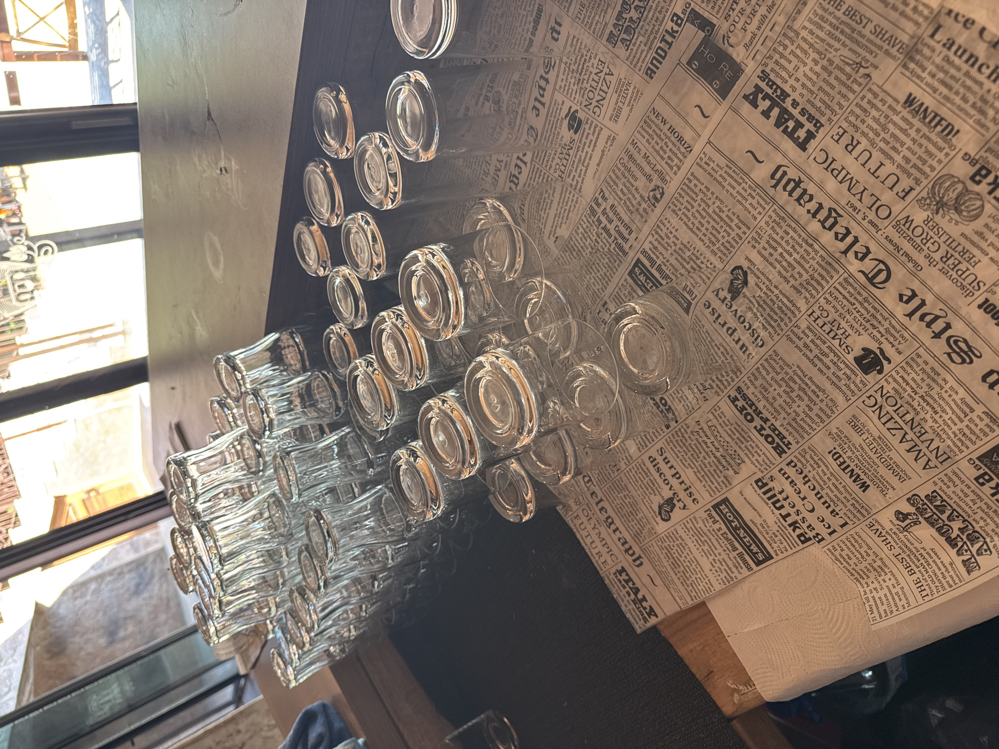 The image shows a bar counter with rows of clean, empty glasses. The glasses are mostly clear and appear to be made of glass, with some reflecting light. Some of the glasses are stacked upright, while others are inverted. Beneath the glasses, there is a surface that appears to be a dark-colored countertop. A newspaper is spread out on the counter beneath some of the glasses, with various headlines and text visible. The newspaper has a sepia tone, suggesting it may be old or intentionally aged. In the background, through a window, outdoor scenery is visible, with sunlight illuminating the area. The overall impression is of a bar or restaurant setting that is prepared for service or has recently finished. There are no people visible in the image. There is no information within the image that suggests the location is in Sredets, Bulgaria.