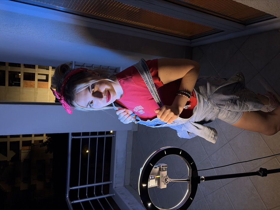 A female individual is depicted posing on an outdoor residential balcony at night in Budva, Montenegro. She has blonde hair styled in a high bun with a pink bandana, and is wearing a red t-shirt, light blue denim overalls, and pink athletic shoes. Multiple beaded bracelets are visible on her right wrist, and she has red lipstick. Her stance is slightly turned towards the camera, with her right hand gripping the strap of her overalls near her shoulder. To the left of the subject, an illuminated ring light with a mounted smartphone stands on a black tripod. The balcony floor consists of light-colored square tiles, and a white metal railing with horizontal bars is present on the left. A white structural column is visible behind the subject. On the right, a large window or glass door with horizontal blinds reflects a cool blue light. The background beyond the balcony reveals dark, indistinct urban buildings, one appearing to be an unfinished concrete structure, under a dark night sky.