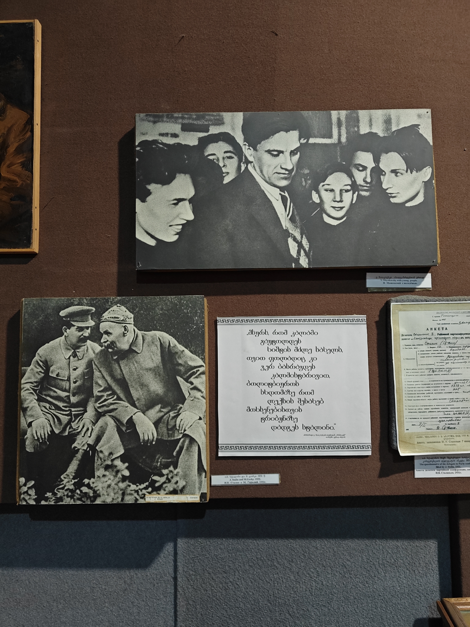 An indoor shot reveals a museum or exhibition display against a textured, dark brown wall. The primary focus is on three historical artifacts: two black and white photographs and two framed documents, each with explanatory captions.
In the upper center, a black and white photograph features several young people, predominantly men, clustered together. One prominent figure, identified by the accompanying caption, is Vladimir Mayakovsky, shown looking left with a slight smile. The caption below reads in Georgian, English, and Russian: "V. Mayakovsky with young people."
Below this, on the left, another black and white photograph shows two men seated outdoors, engaged in conversation. One man, on the left, wears a military-style uniform with a cap, a mustache, and a pipe. The man on the right, wearing a cap and a heavy coat, also has a prominent mustache and is gesturing with his hands. The caption identifies them as "J. Stalin and M. Gorky. 1932."
To the right of the Stalin and Gorky photograph, a white sheet of paper displays Georgian text, likely a poem or literary excerpt, framed by a decorative border. A smaller caption below confirms it is "An excerpt from V. Mayakovsky's poem."
Further to the right, an older, aged document titled "АНКЕТА" (Questionnaire) is displayed. It features Cyrillic text, with handwritten entries visible. The caption below translates to "The questionnaire of the delegate to Party conference, filled by J. Stalin. 1931."
Partially visible on the far left edge of the image is a section of a framed painting. The overall impression is a curated display of historical figures and documents, possibly related to Soviet-era history or Georgian literary and political figures.