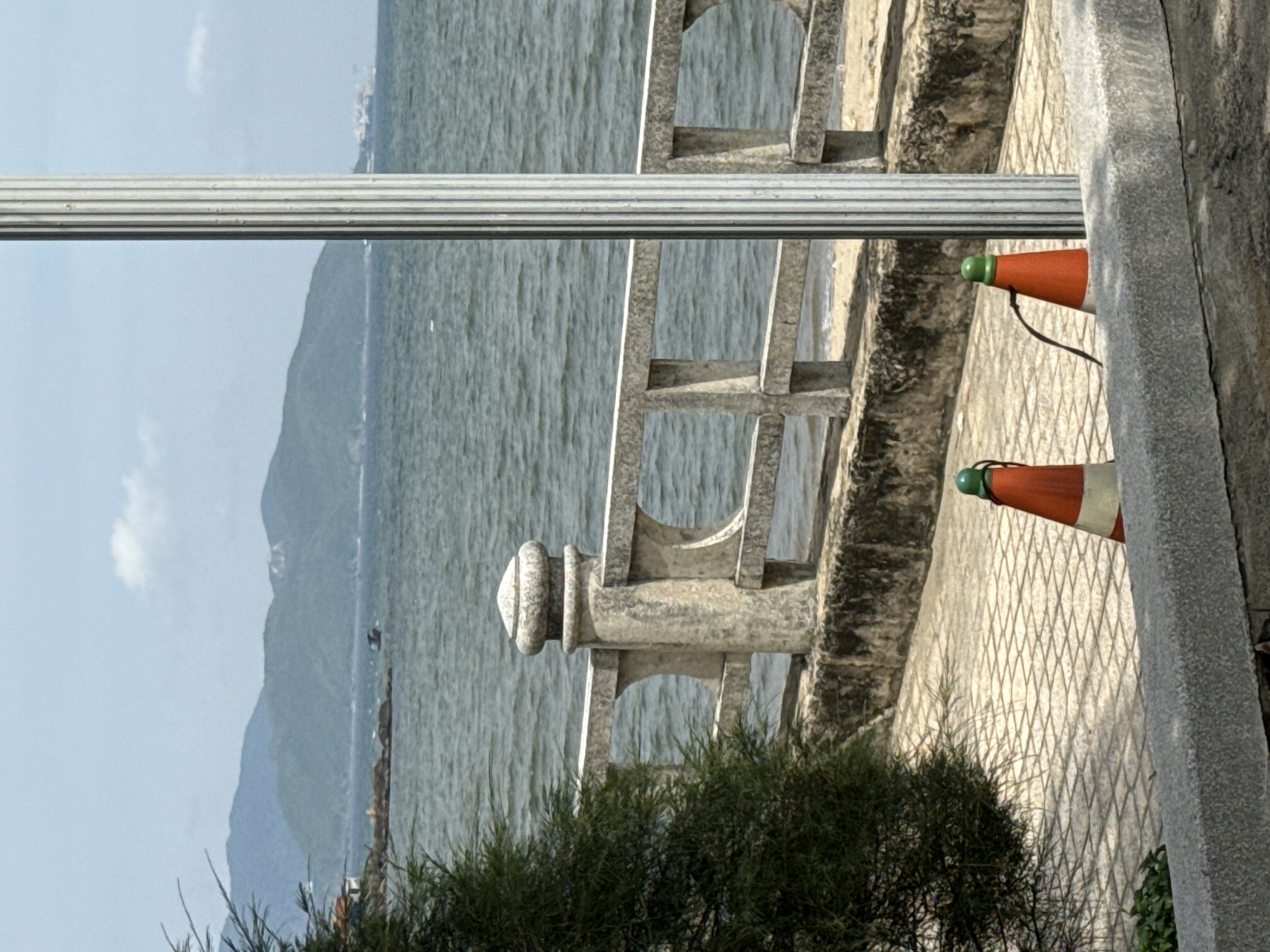 A vertical, fluted metal pole stands in the foreground on a paved walkway beside a body of water. To the left of the pole, a dense, dark green tree with fine needles partially obscures a weathered, light-colored concrete railing with decorative arches. Two orange traffic cones with white bands and green tops are positioned on the walkway near the pole, with a black wire connecting them. The walkway is paved with light-colored, diamond-patterned tiles. Beyond the railing, the water is a muted green with ripples, and a sandy beach is visible in the distance. In the background, several large, rounded mountains with a hazy, blue-green hue rise from the water. The sky is a pale blue with scattered white clouds. The scene appears to be a coastal area.
