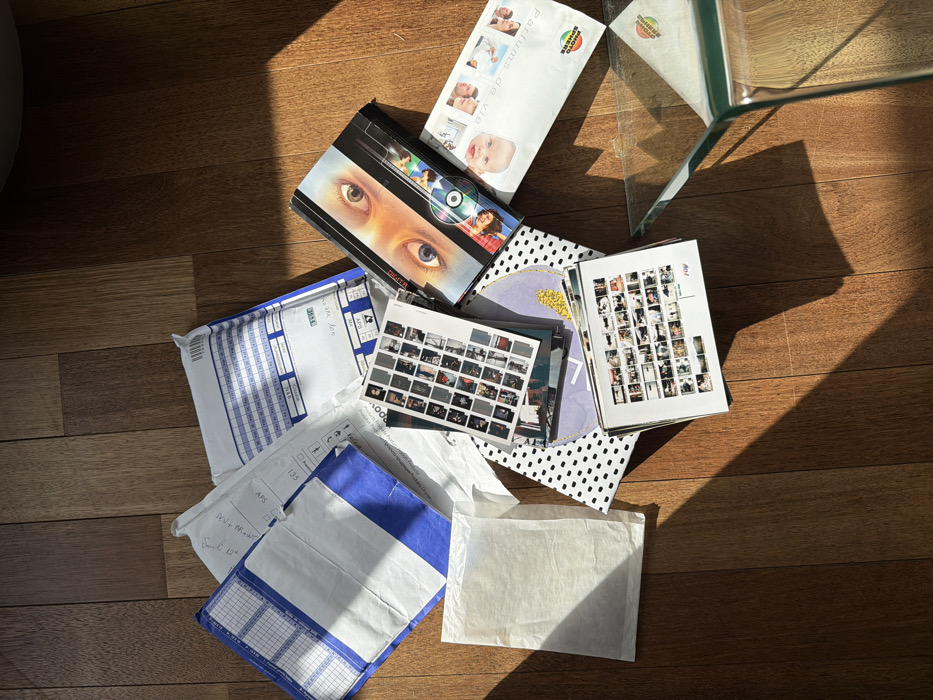 An overhead view captures a collection of mail, packages, and photographic materials spread across a light-to-medium brown wooden floor in an interior setting within Champs-sur-Marne, France. Direct sunlight casts strong shadows from the upper right toward the lower left, illuminating approximately half of the scene.
The arrangement includes two white padded envelopes with distinct blue striped patterns and grid-lined labels, likely from a postal service, one appearing partially opened. A smaller, thin white paper envelope lies to the left. Central to the composition is a stack of various photographic prints and contact sheets; one clearly visible sheet displays numerous small, square, predominantly black and white images arranged in a grid. Another prominent item is a book or DVD case titled "DIGITAL," featuring a close-up image of two human eyes (one brown, one blue) and a reflective disc. Adjacent to this is a white envelope from "PHOTO SPHERE," adorned with images of babies and the text "PARFUMS de vie." A black and white polka-dotted sheet of paper or board underlies some of these items. In the bottom right corner, the clear, angular edge of a glass table is visible, reflecting ambient light. No individuals are present in the scene.