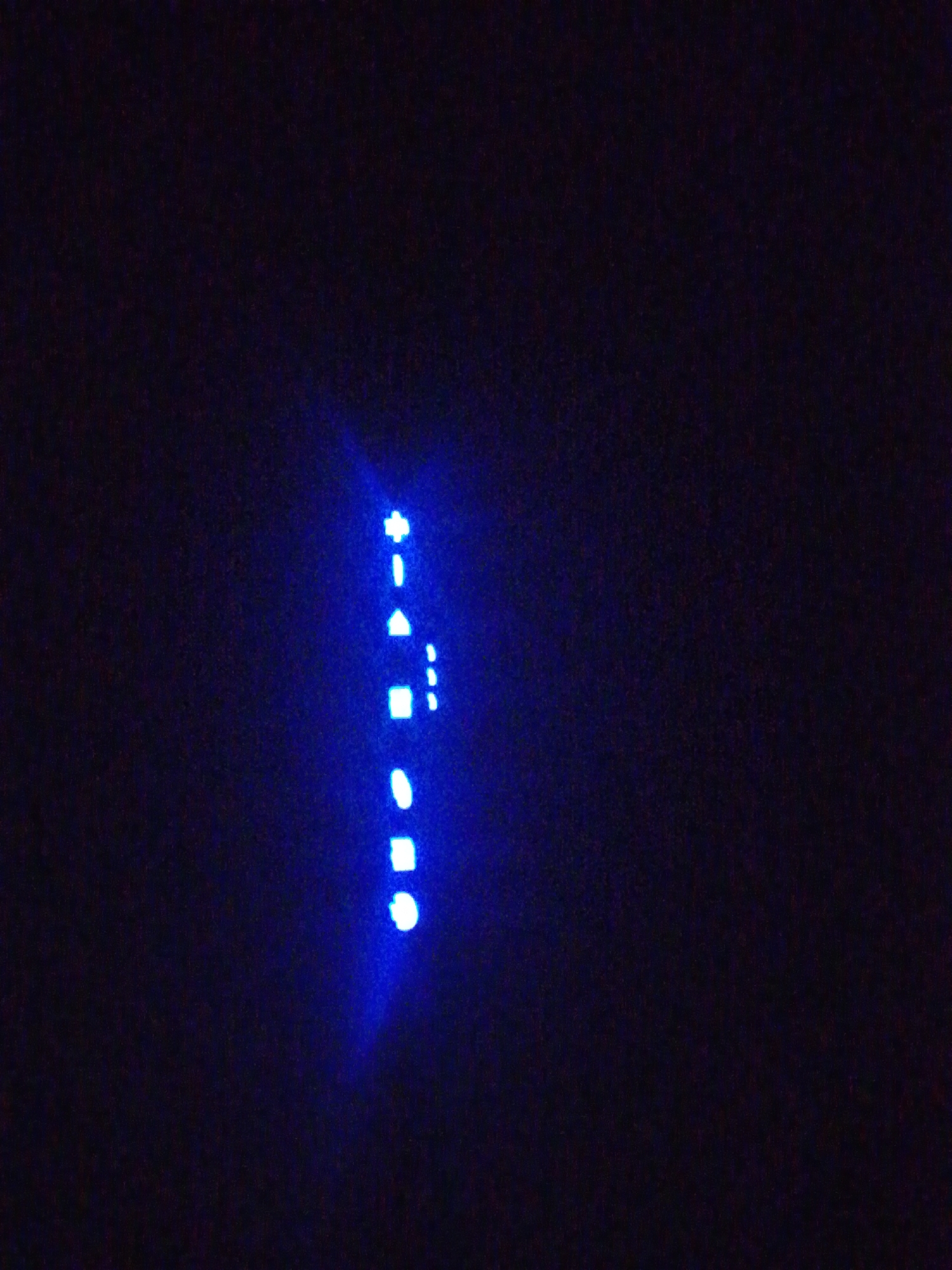 The image displays a dark, low-light environment dominated by blue illumination. A vertical series of distinct blue light forms is positioned on the right side of the frame. From top to bottom, these shapes include a cross or plus sign, a short horizontal dash, an upward-pointing triangle, a square, a small oval or circular shape, another square, and a brighter, larger oval or circular shape at the bottom. To the right of the triangle and the first square, several smaller, less defined blue dots or dashes are visible. The background is a uniform dark blue-black, indicating the absence of ambient light, and the blue light sources appear to emit a soft glow into the surrounding darkness. No discernible details regarding people, specific objects beyond the lights, actions, interactions, or the city of Keffi, Nigeria, are present within the visual information provided.