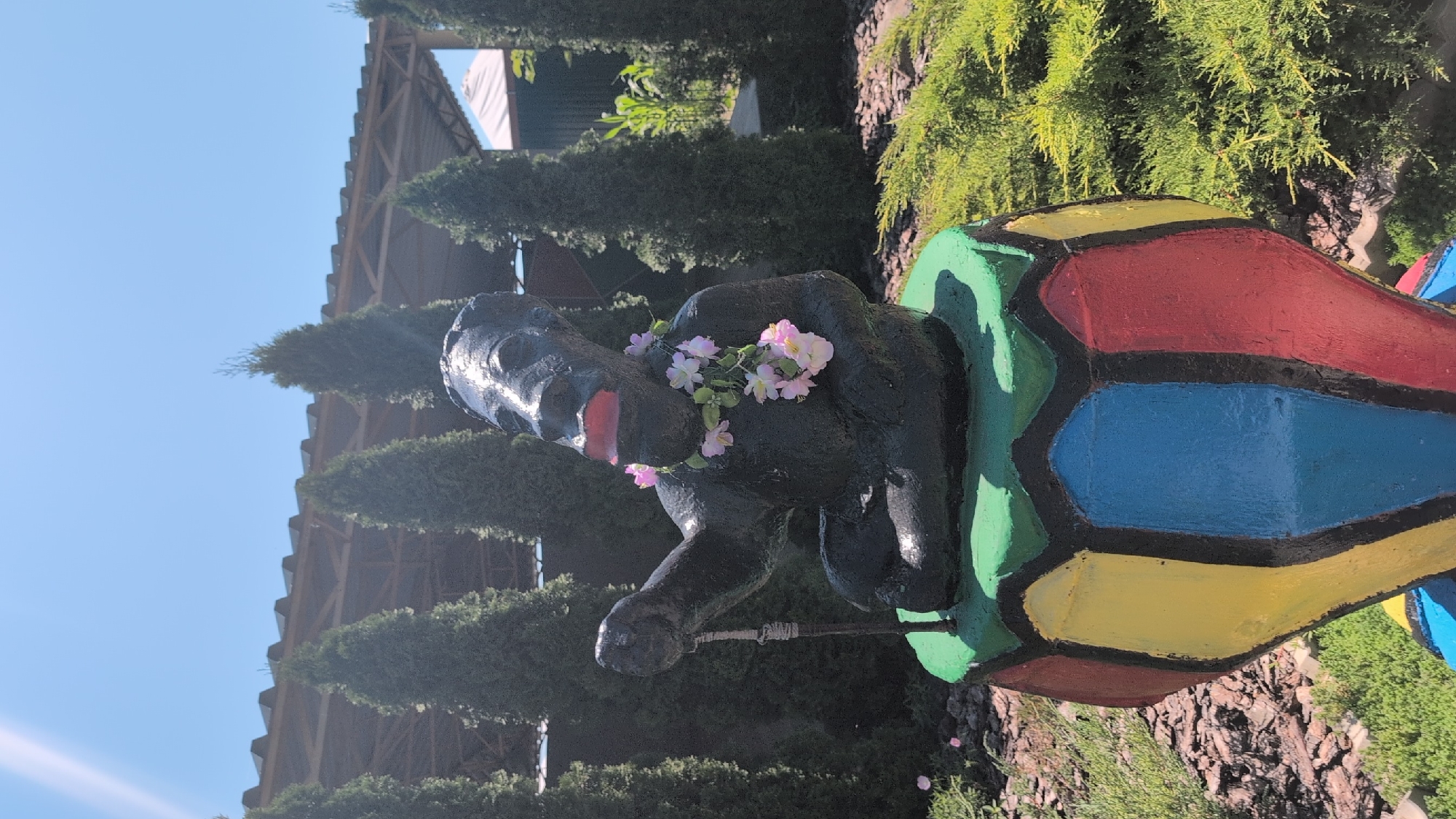 A dark-colored statue of a seated figure wearing a necklace of pink flowers sits atop a brightly painted drum. The statue has a dark face with prominent lips painted red, and it holds a staff in its right hand. The drum is decorated with sections of yellow, blue, and red paint, outlined in black. The statue is outdoors, with a clear blue sky and a wooden roof structure visible in the background. Several tall, dark green coniferous trees line the background, and lush green bushes are in the foreground and to the right. The ground around the statue is covered with bark mulch. This is a sculpture located in the Izmail City Park in Izmail, Ukraine.