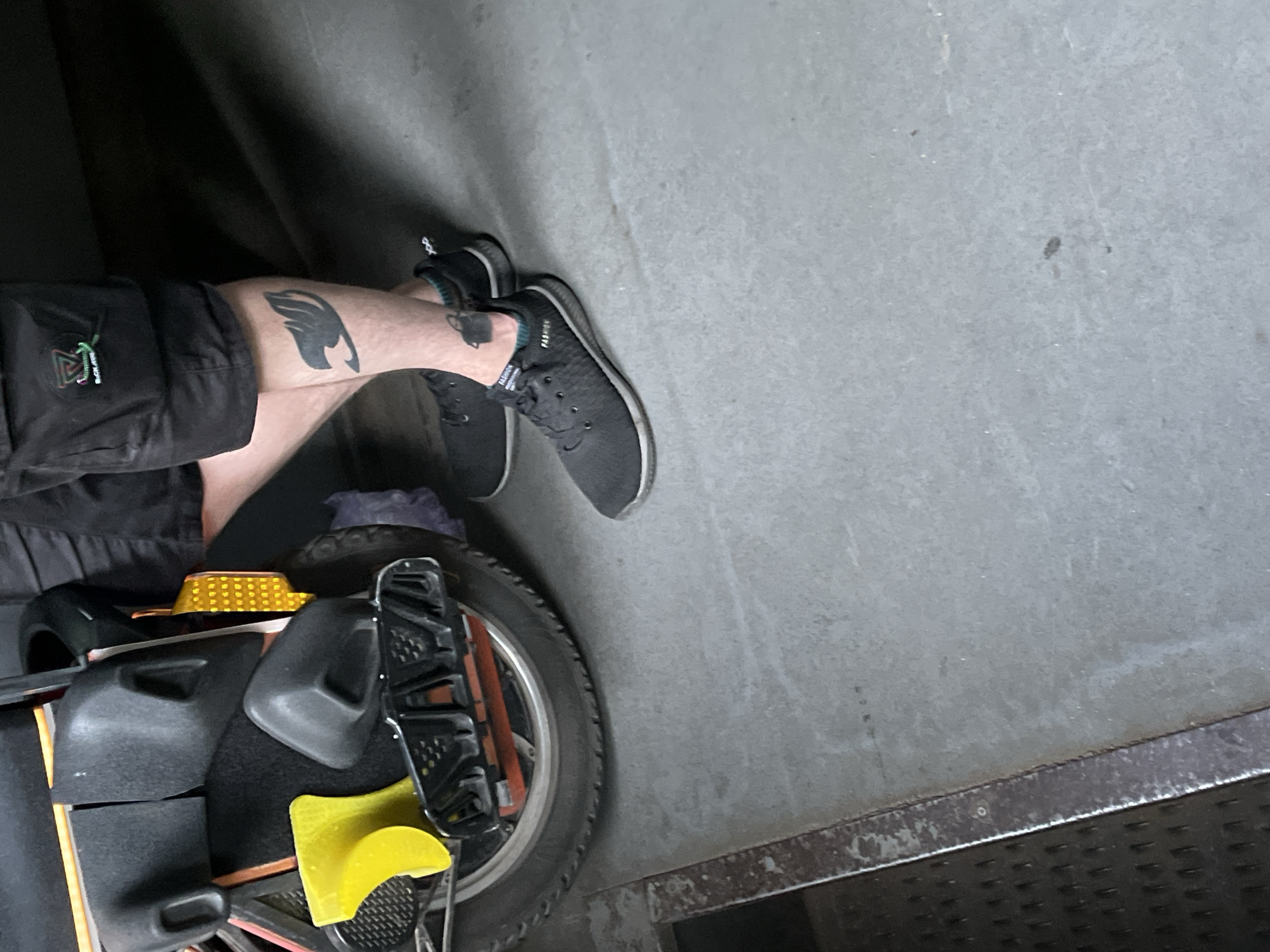 The media file shows a person's legs and feet resting on what appears to be an electric unicycle. The person is wearing black shorts with a small logo containing geometric shapes in green, white, and red, and black athletic shoes. A prominent black tattoo of a wing is visible on the person's left thigh. The unicycle is black and grey with yellow accents and a black tire. The background is a textured grey concrete surface, possibly an urban or industrial setting. The lighting suggests it might be daytime, but the environment appears to be somewhat shaded. There is no clear indication of the weather or specific emotions. The visible text on the shoe reads "FASHION". The image provides a close-up view of the legs and the unicycle, with the background mostly out of focus.