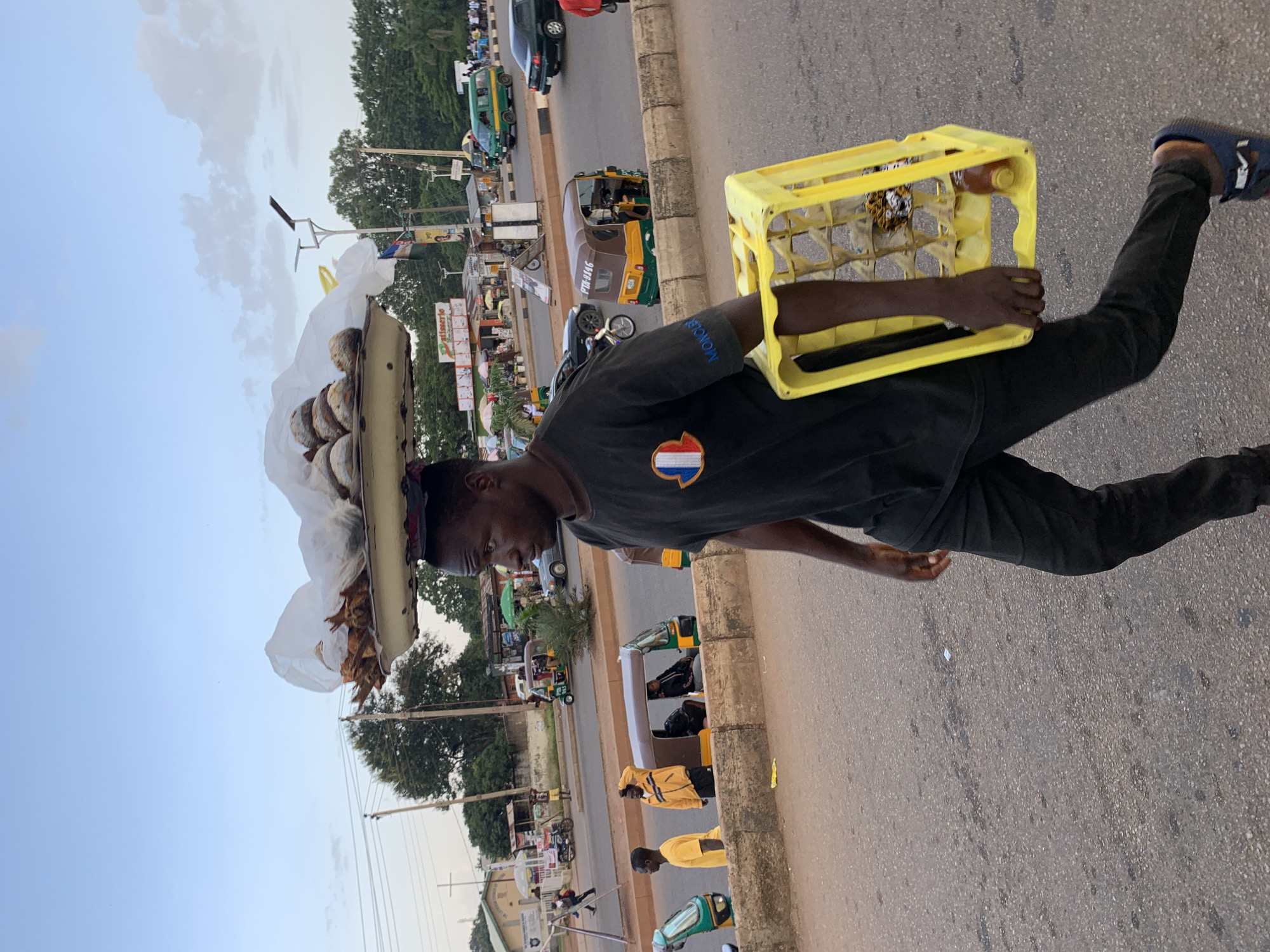 A man walks down a street in Jos, Nigeria, carrying a tray of food items on his head and a yellow plastic crate in his hand. He is wearing a black t-shirt with a small French flag logo and black pants. The food on the tray appears to be dried fish and other items covered with plastic. The crate contains bottles, one of which is visible and appears to be a drink. In the background, there are several vehicles, including a green van, a black sedan, and a few auto-rickshaws. Buildings and trees line the street, and a cloudy sky is visible. The street is paved, and there is a concrete barrier separating the traffic lanes from the pedestrian area. The overall scene depicts a bustling street in an urban environment.