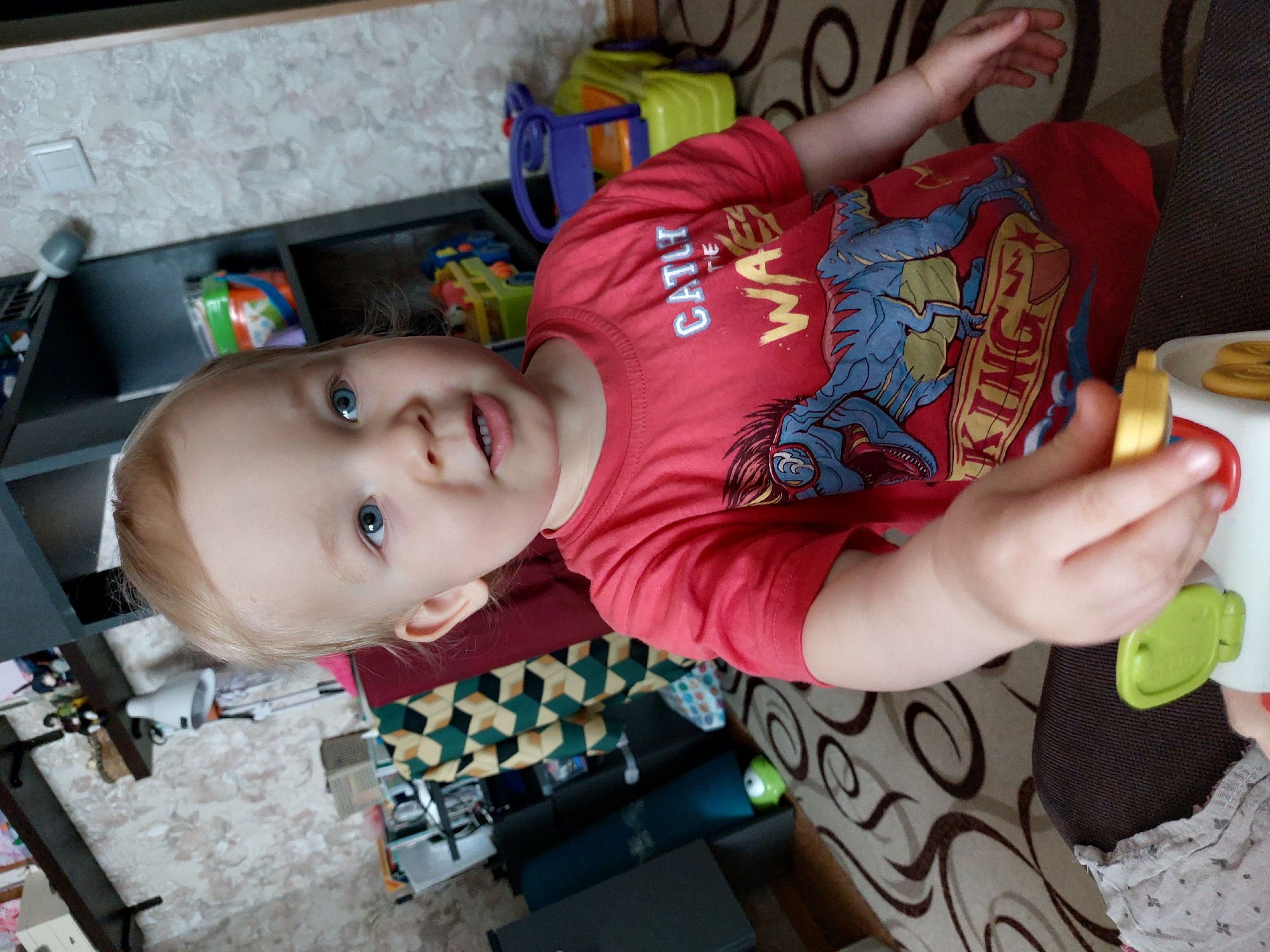 A fair-skinned toddler with blonde hair and blue eyes, wearing a red t-shirt with a dinosaur print, is in a high chair, holding a toy. The toddler is indoors, likely in a home. In the background, there is a shelf with toys and other items. There is also a patterned rug on the floor. No information is available to confirm it is in Chaikovsky, Russia.