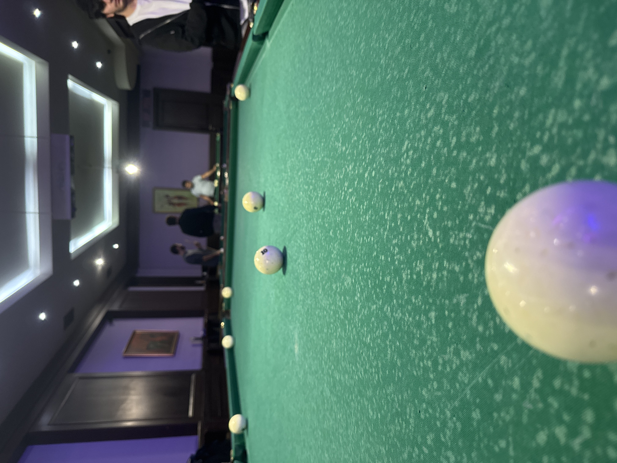 The image shows a billiard table in what appears to be a billiard hall. The table is covered in green felt, and several white billiard balls are scattered across the surface. One ball, prominently positioned in the foreground, is white with a numbered sticker. People are vaguely visible in the background, suggesting an active environment. The interior decor includes framed artwork and ceiling lighting. Given the request to incorporate location, the scene is possibly in a billiard hall within the city of Yerevan, Armenia. No specific actions or interactions are definitively visible beyond the presence of people in the background.