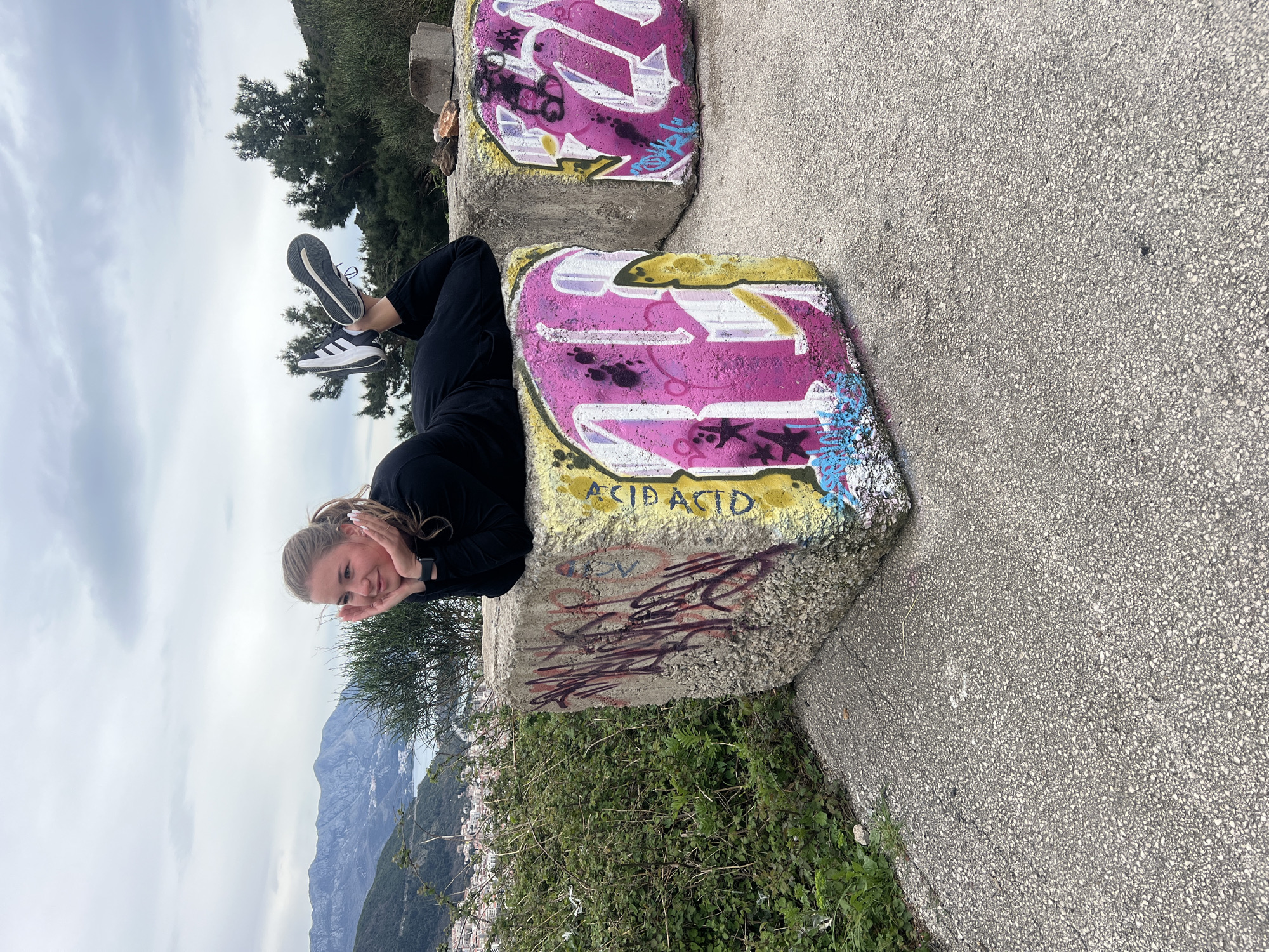 A girl lies between two concrete blocks, her head resting on her hands, with her legs and feet raised in the air. She has long blonde hair. She wears black leggings and a black sweater, along with white Adidas sneakers with black stripes. The blocks are covered in graffiti, with the words "ACID ACID" visible on the lower block. In the background, there is a view of mountains and the city of Budva, Montenegro. The sky is overcast. A paved surface is visible on the right, with green vegetation below the blocks.