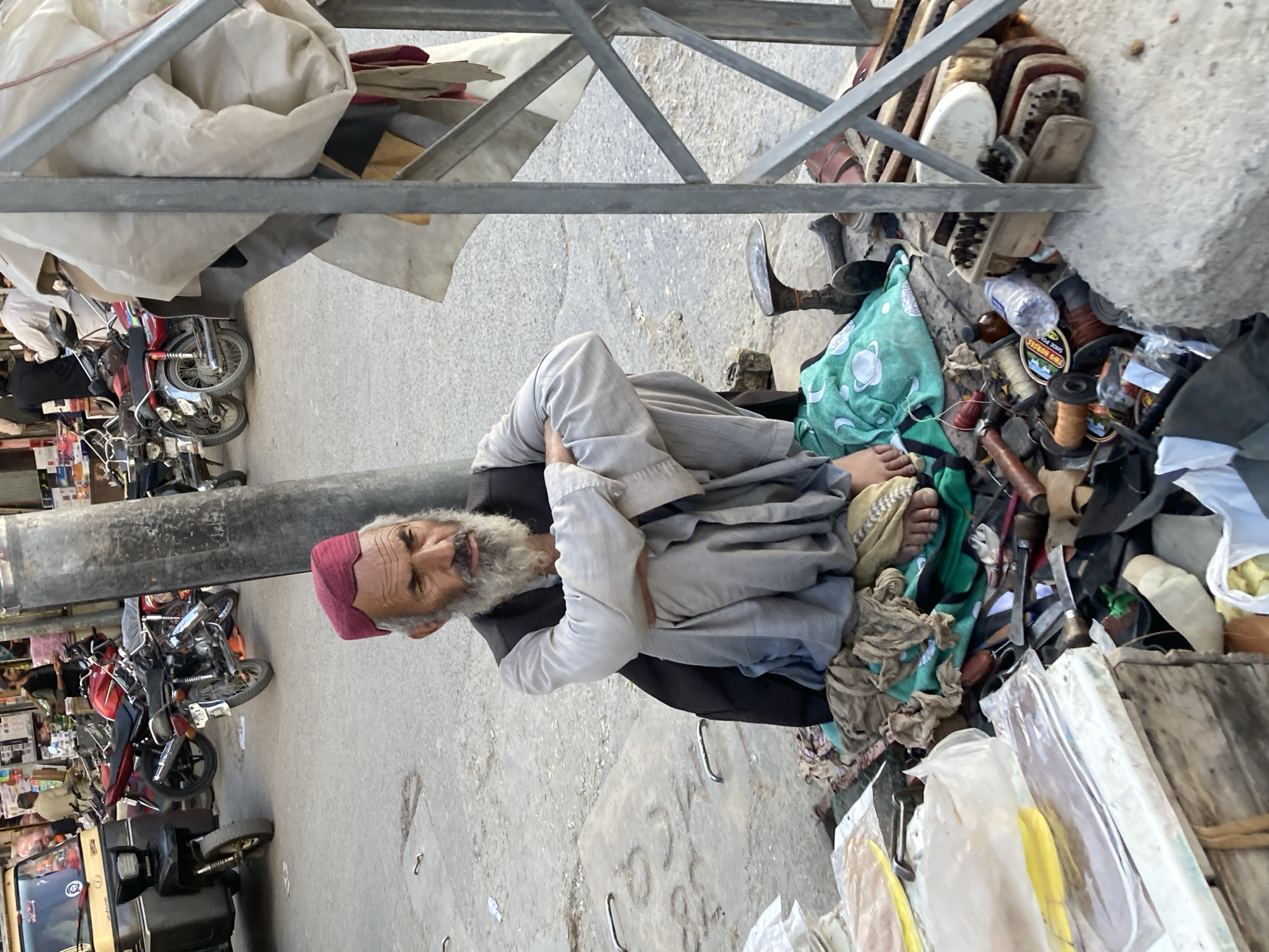 The image shows an elderly man with a white beard and a red cap sitting on the ground near a pile of objects that includes shoes, leather goods, and sewing supplies. He is wearing light-colored clothing and appears to be resting or working among his possessions. A metal structure is visible overhead, and in the background, there's a street scene with motorcycles, a tuk-tuk, and buildings. The location appears to be a street in Quetta City Tehsil, Pakistan.